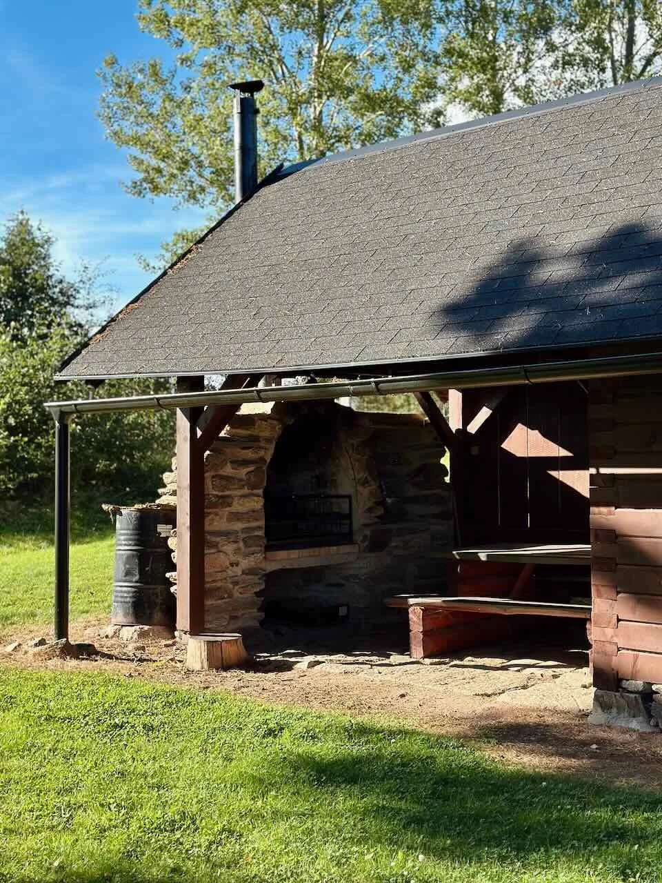 A rustic outdoor shelter with a stone and wood structure, a pitched shingle roof, and a nearby metal barrel, surrounded by green grass and trees under a clear blue sky.