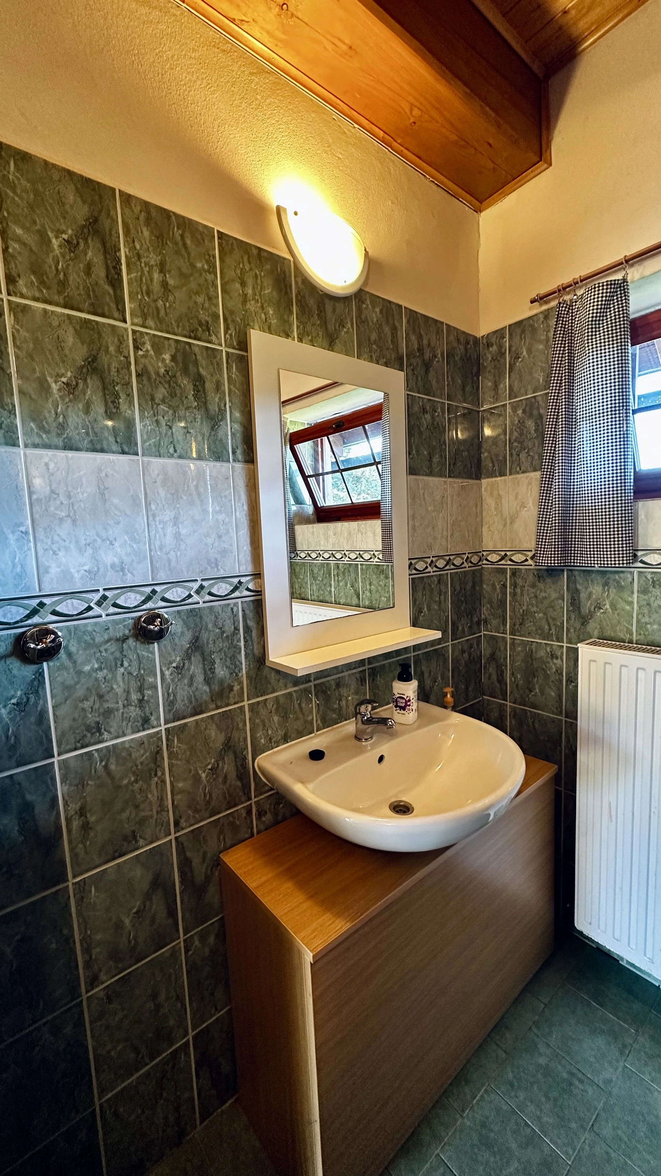 Bathroom with green and beige tiled walls, wooden ceiling, small sink on a wooden vanity, mirror above the sink, window with checkered curtain, radiator, and wall-mounted hooks.