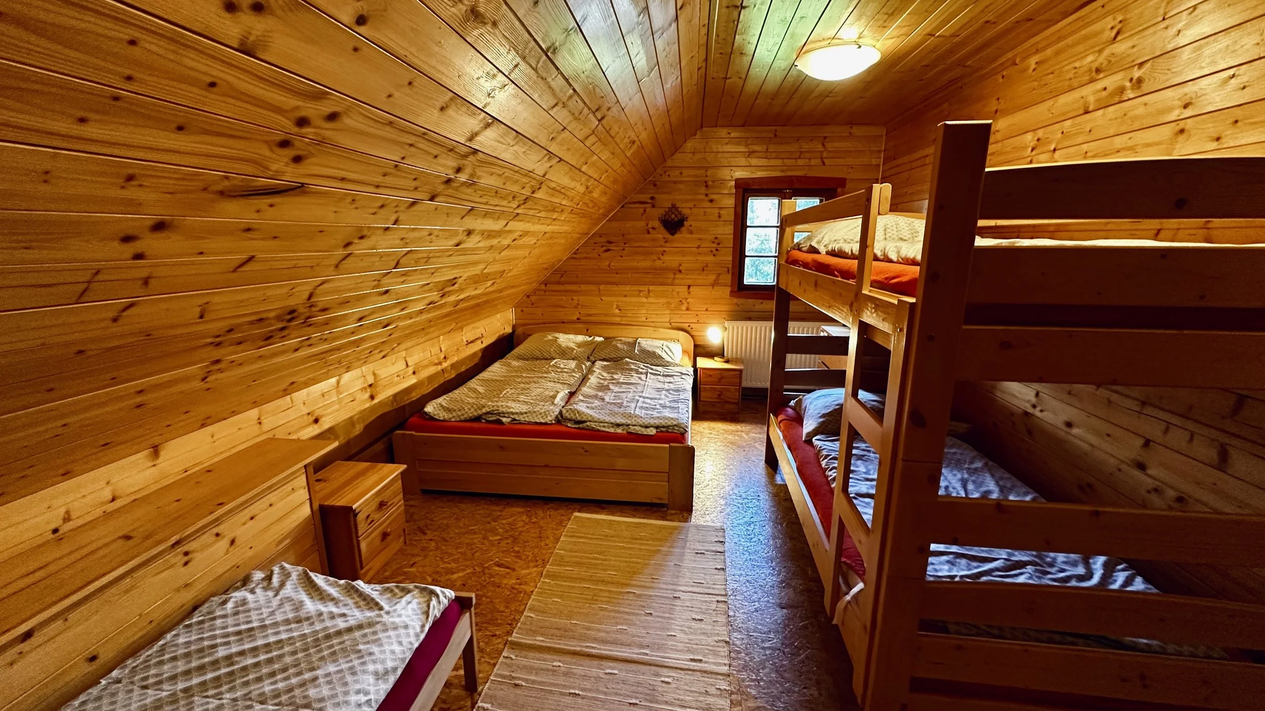 Wood-paneled attic bedroom with a double bed, bunk beds, a nightstand with a lamp, and a small window.