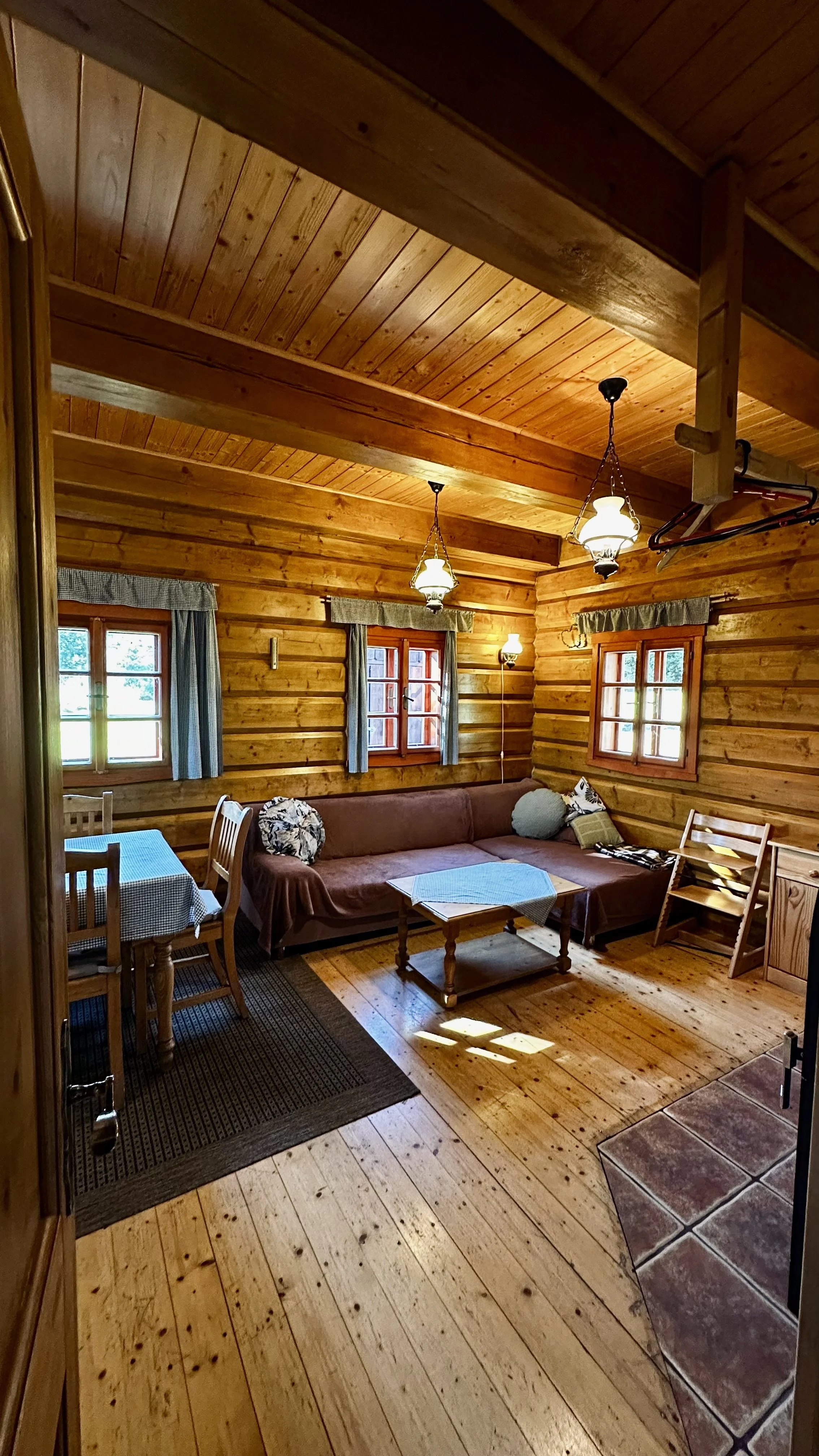 A cozy wood-paneled living room with a brown sofa, small wooden coffee table, two side tables, three small windows with curtains, a dining table with chairs, a hanging light fixture, and wooden flooring.