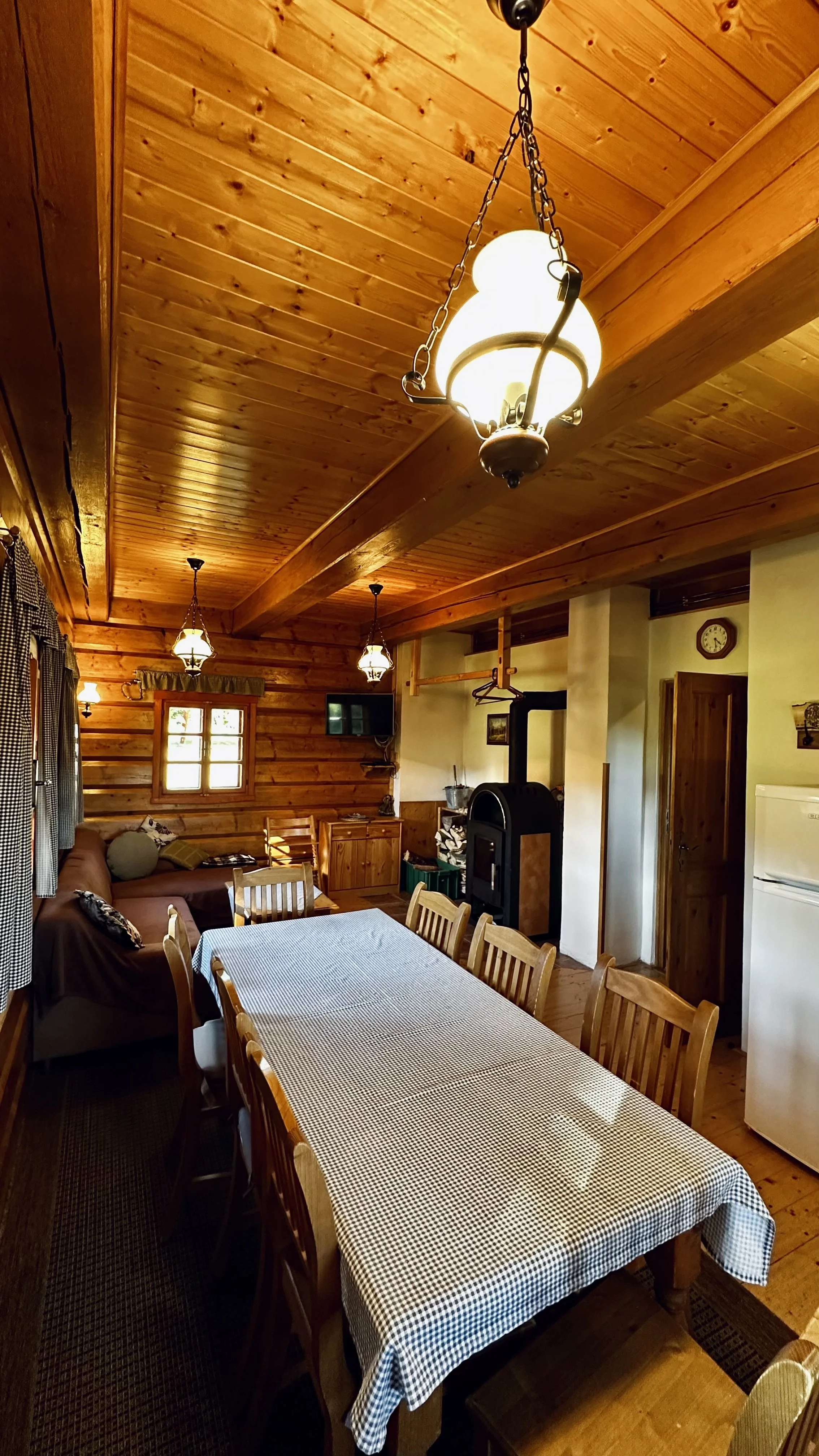 Interior of a rustic wooden cabin dining and living area with a long checkered table, wooden chairs, a sofa, and cabin decor including a wood stove, small TV, and vintage wall clock.