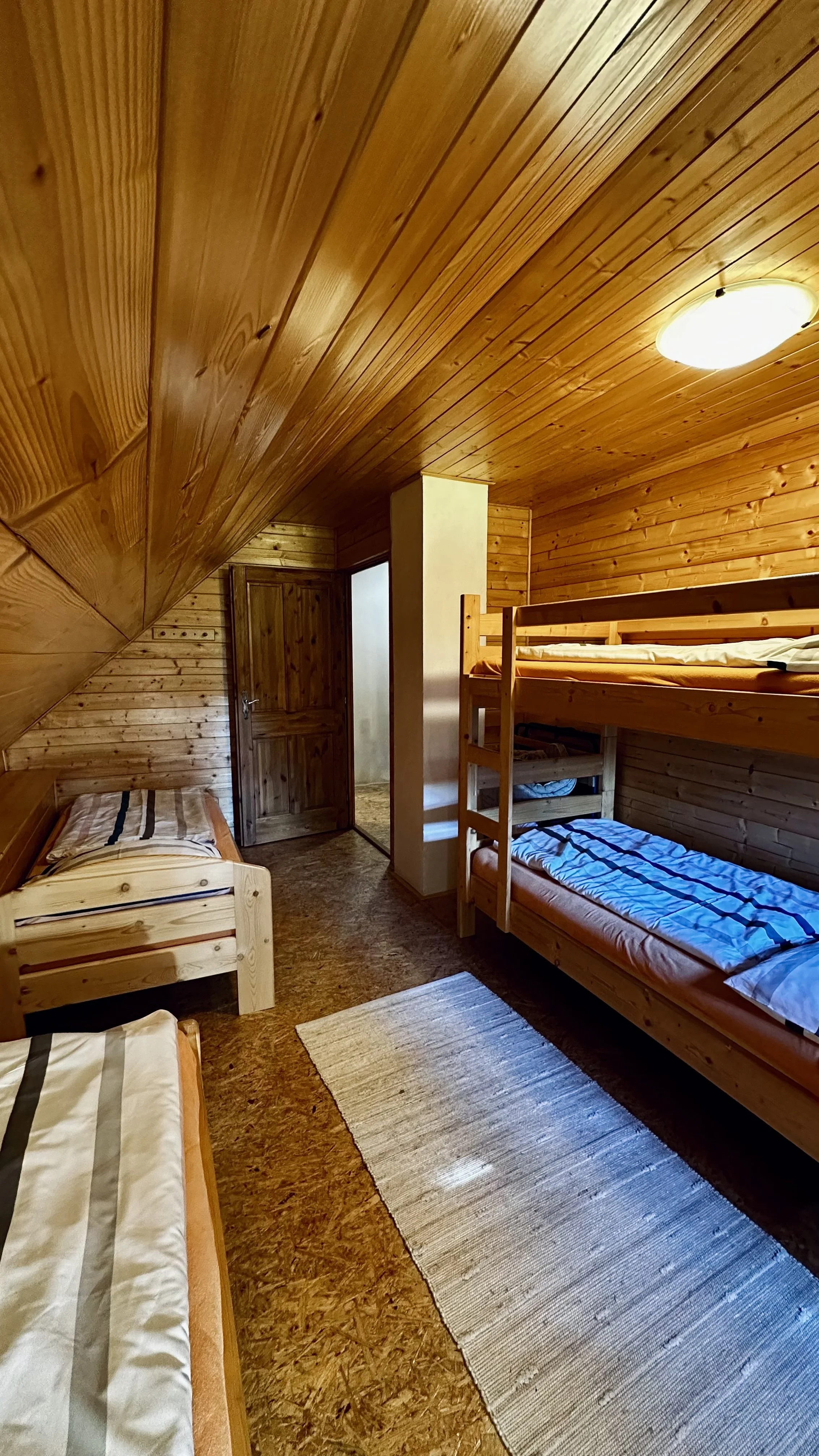 A cozy bedroom with wooden paneling on the walls and ceiling, featuring two single beds, one with a striped blanket and one with a blue striped blanket, a small rug on the floor, and a wooden door.