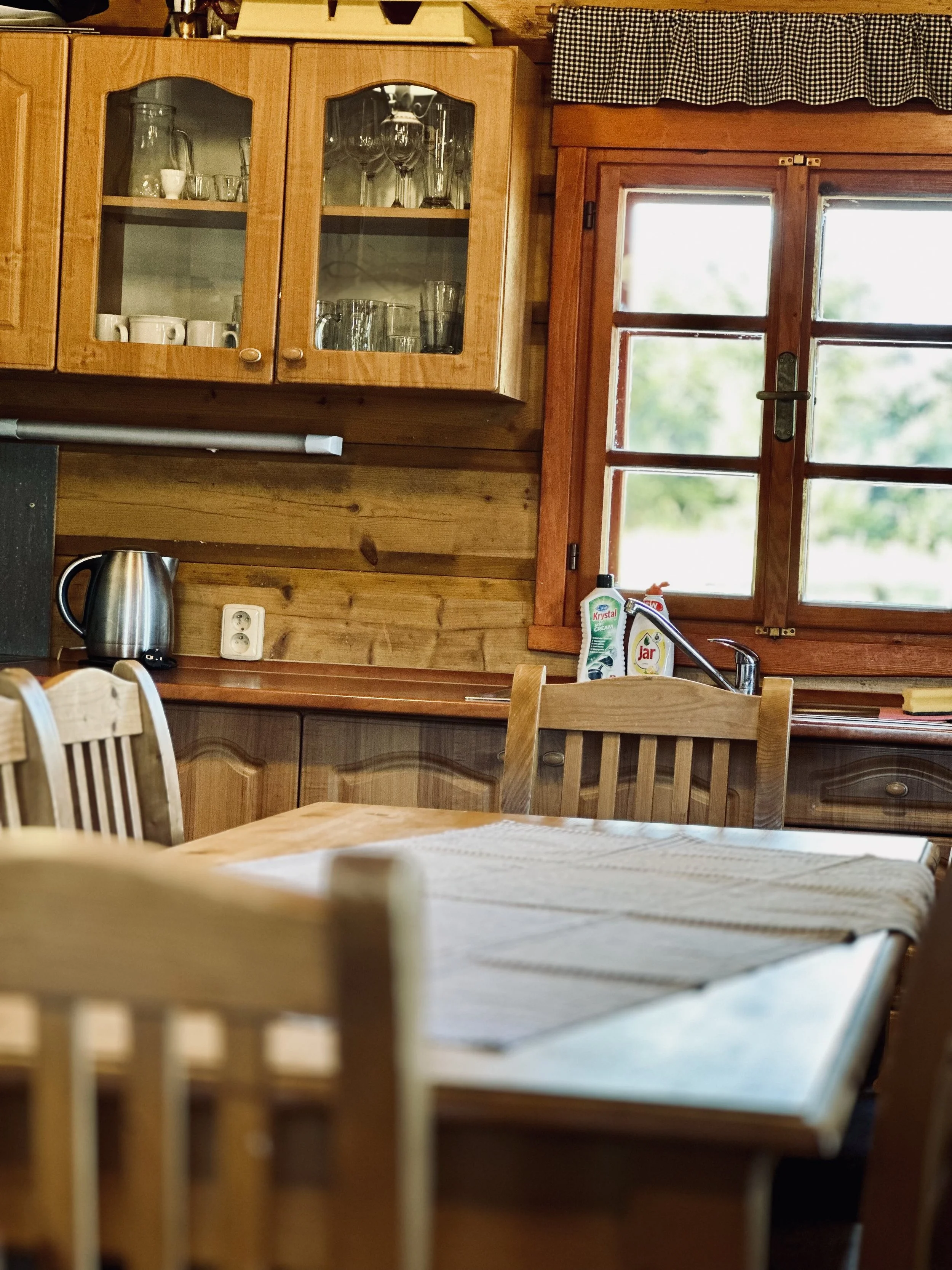 A cozy rustic kitchen with wooden cabinets, a dining table with chairs, a window with a wooden frame, and kitchen items like a kettle, dish soap, and a drying rack.