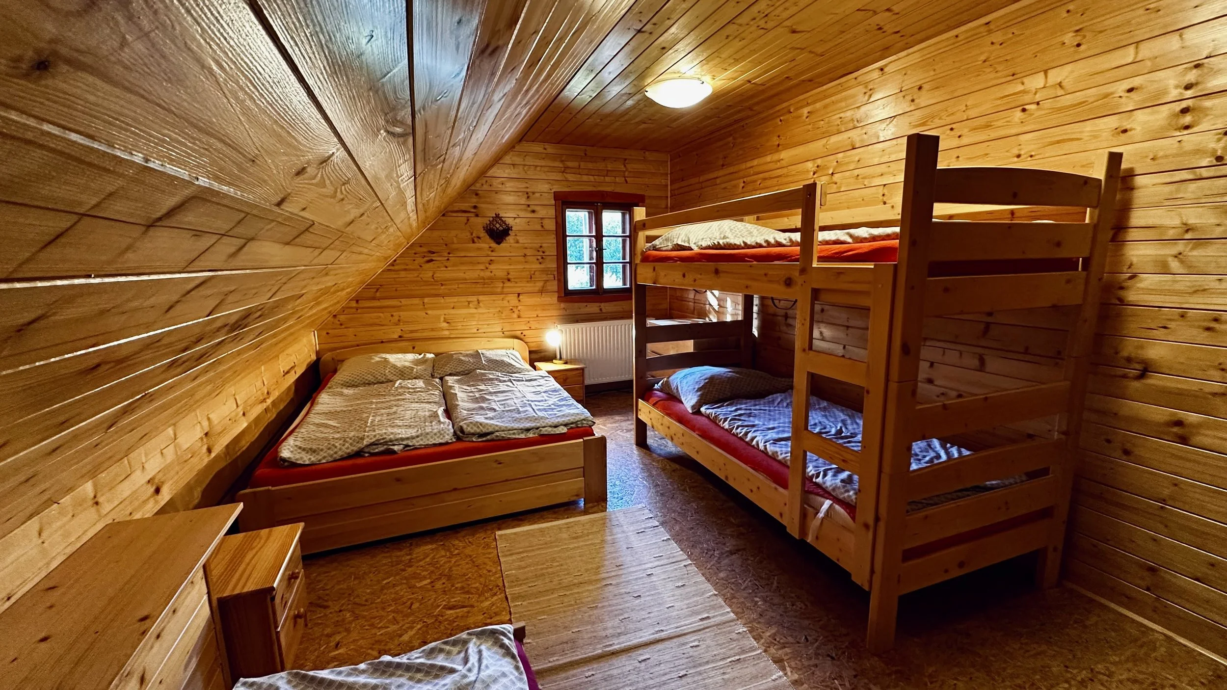 A cozy bedroom with wooden walls and ceiling, featuring a double bed, a bunk bed, a small window, a nightstand with a lamp, and a small dresser.