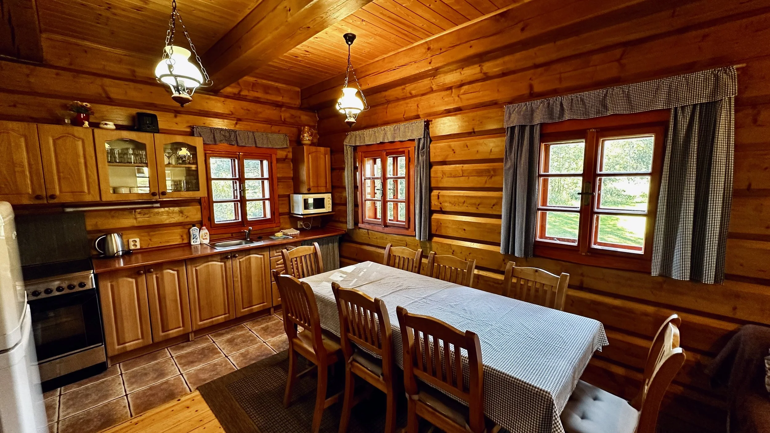 Rustic kitchen with wooden walls, cabinets, and ceiling, featuring three windows with checkered curtains, a dining table with chairs, and kitchen appliances like a stove, microwave, and electric kettle.