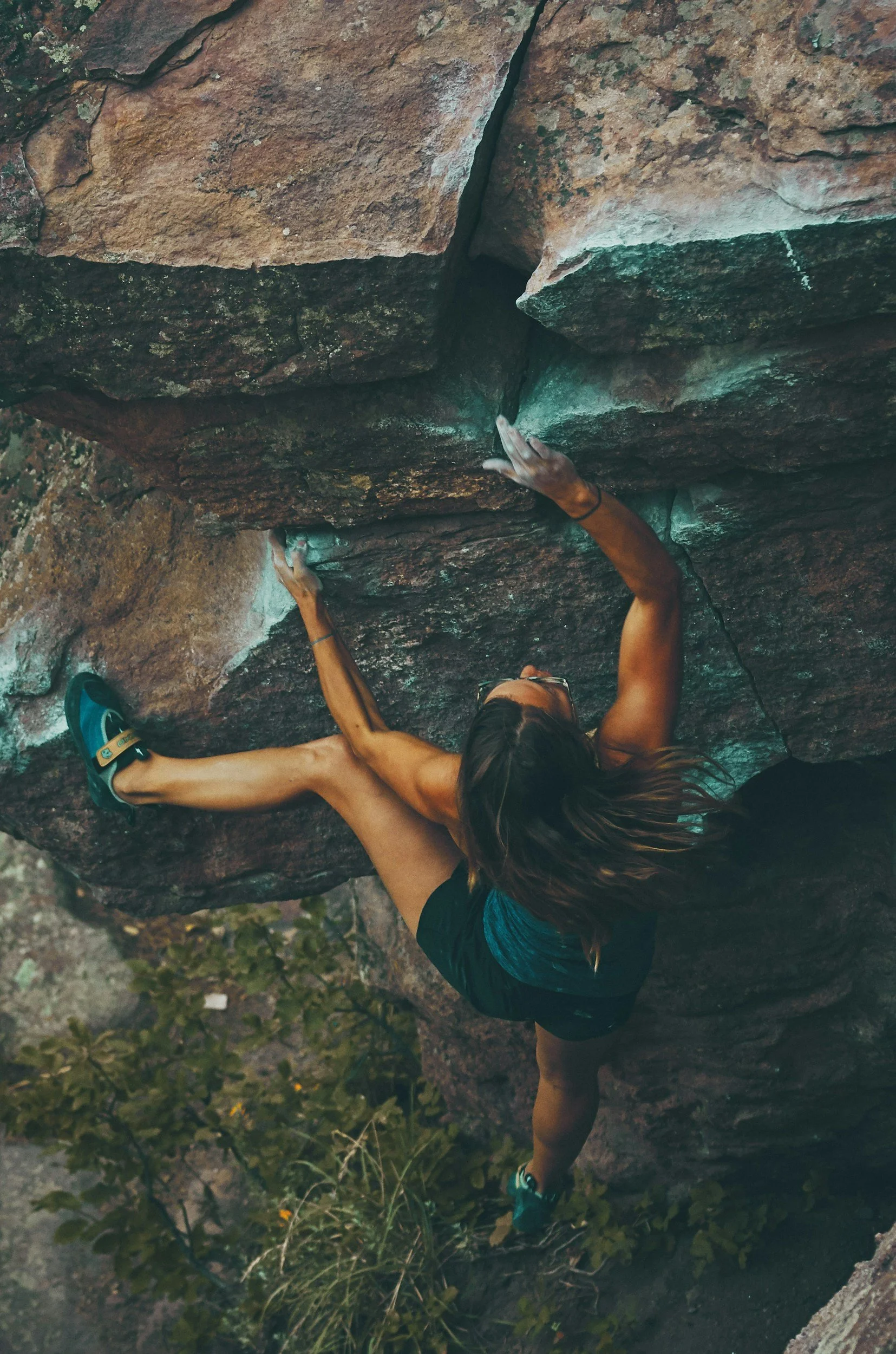 A woman is rock climbing on a vertical outdoor cliff face with greenery below.