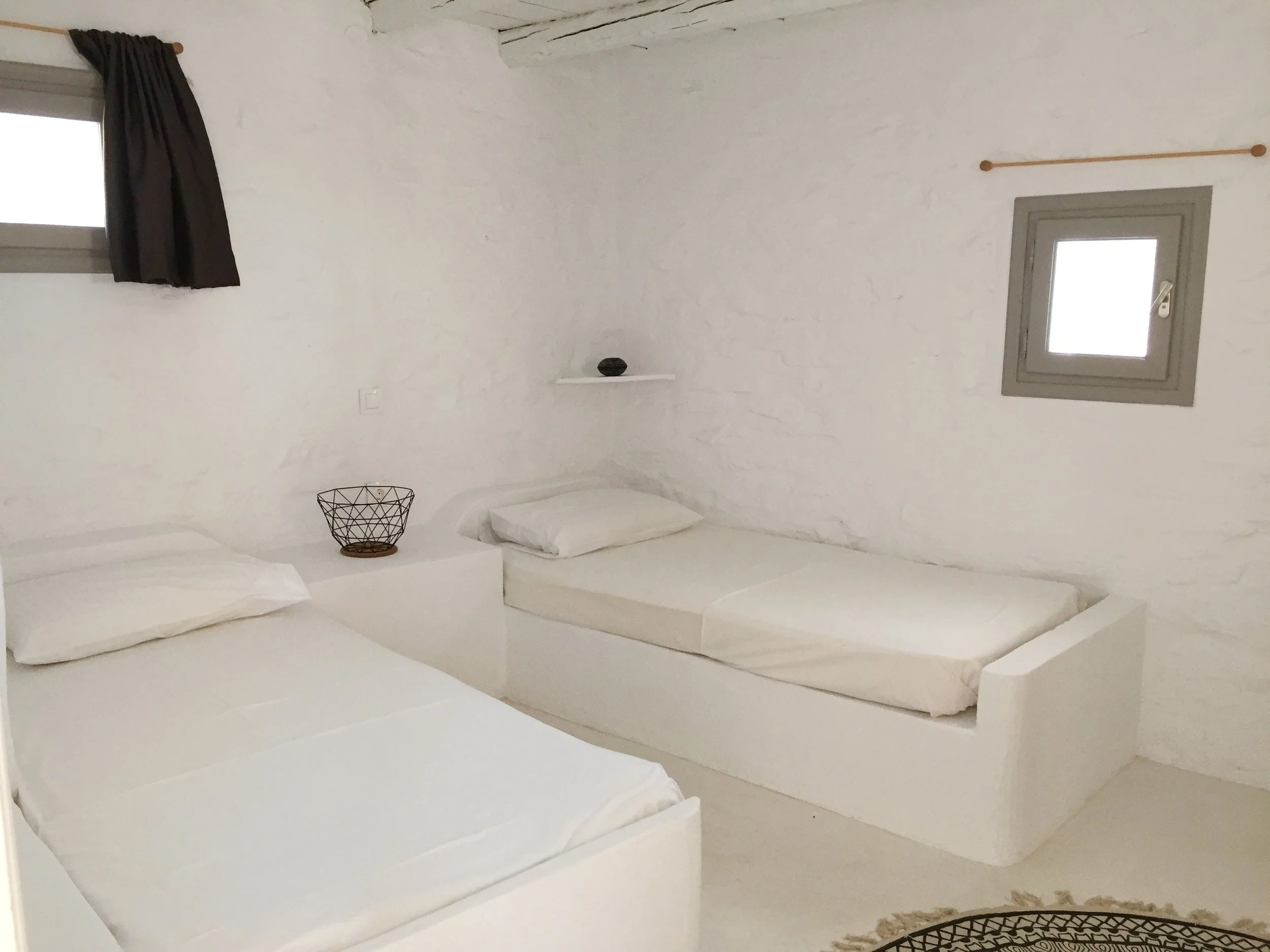 Two neatly made beds with white sheets and folded towels on top, in a minimalist bedroom with white walls, a wooden framed mirror, a black chair, and an air conditioning unit.