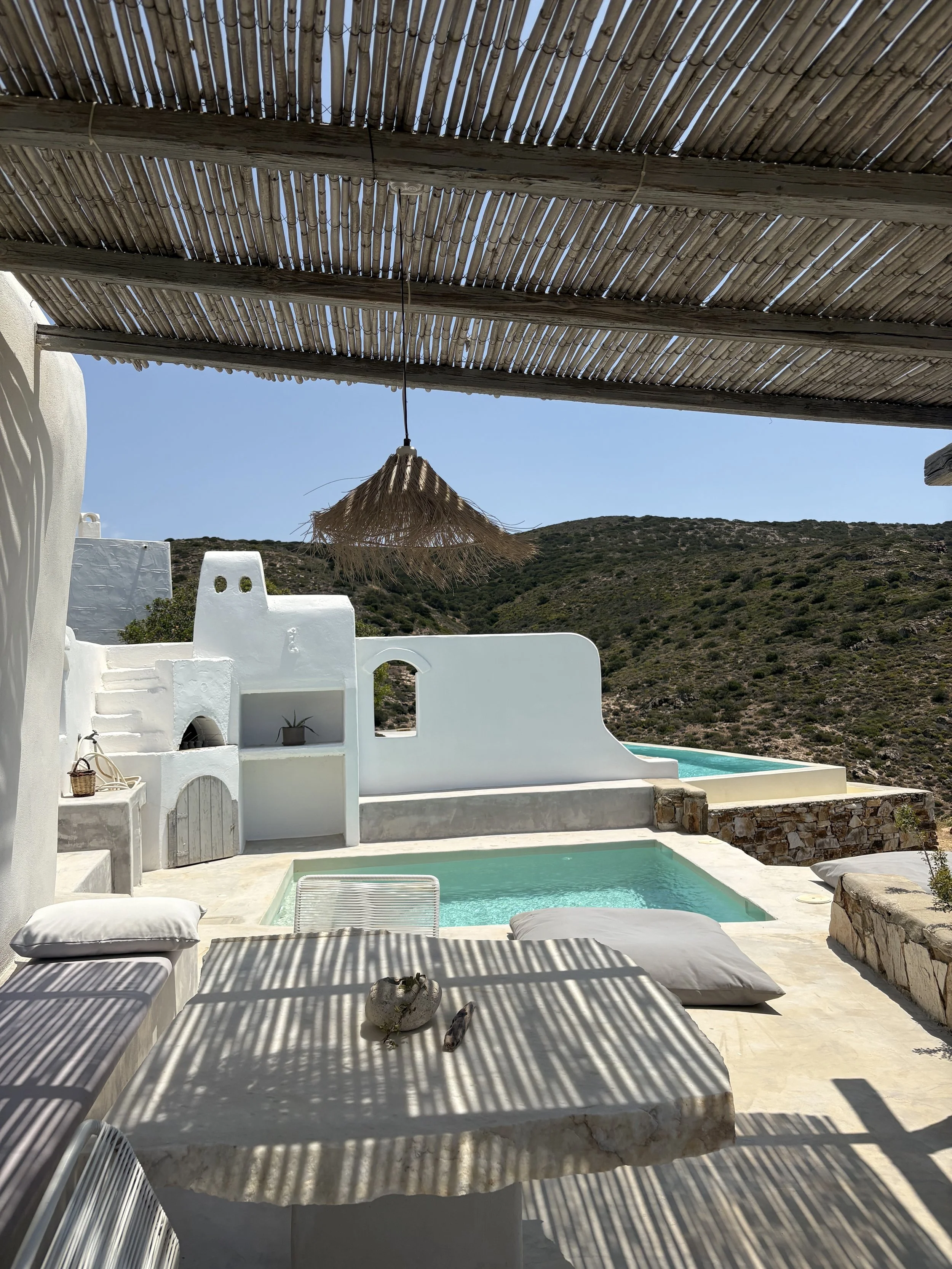 A sunny outdoor patio with a small swimming pool, white stucco walls, and a mountain landscape in the background. There is a shaded area with a bamboo roof, a thatched pendant light, and a table with cushions, shadows cast by the bamboo roof, and a r