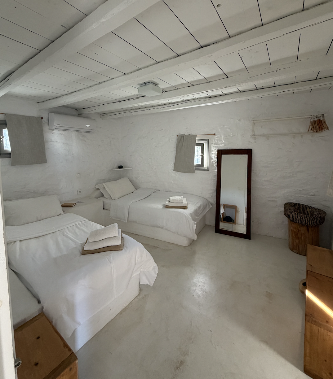 Two neatly made beds with white sheets and folded towels on top, in a minimalist bedroom with white walls, a wooden framed mirror, a black chair, and an air conditioning unit.