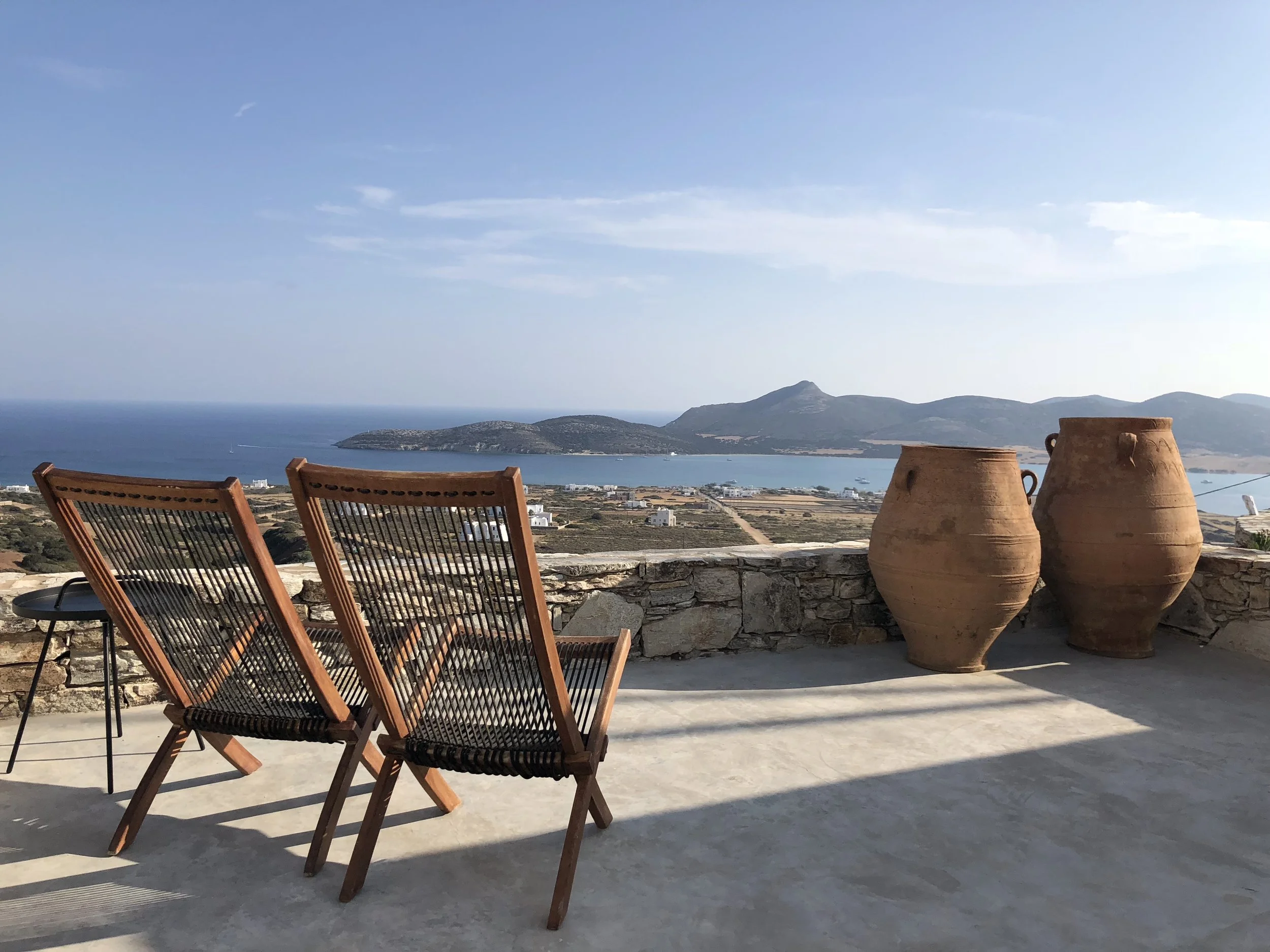 View from a terrace overlooking a coastal landscape with two wooden chairs and two large clay pots in the foreground.