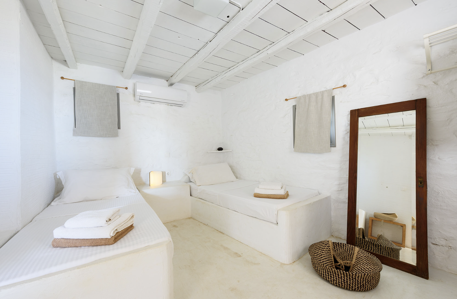Two neatly made beds with white sheets and folded towels on top, in a minimalist bedroom with white walls, a wooden framed mirror, a black chair, and an air conditioning unit.