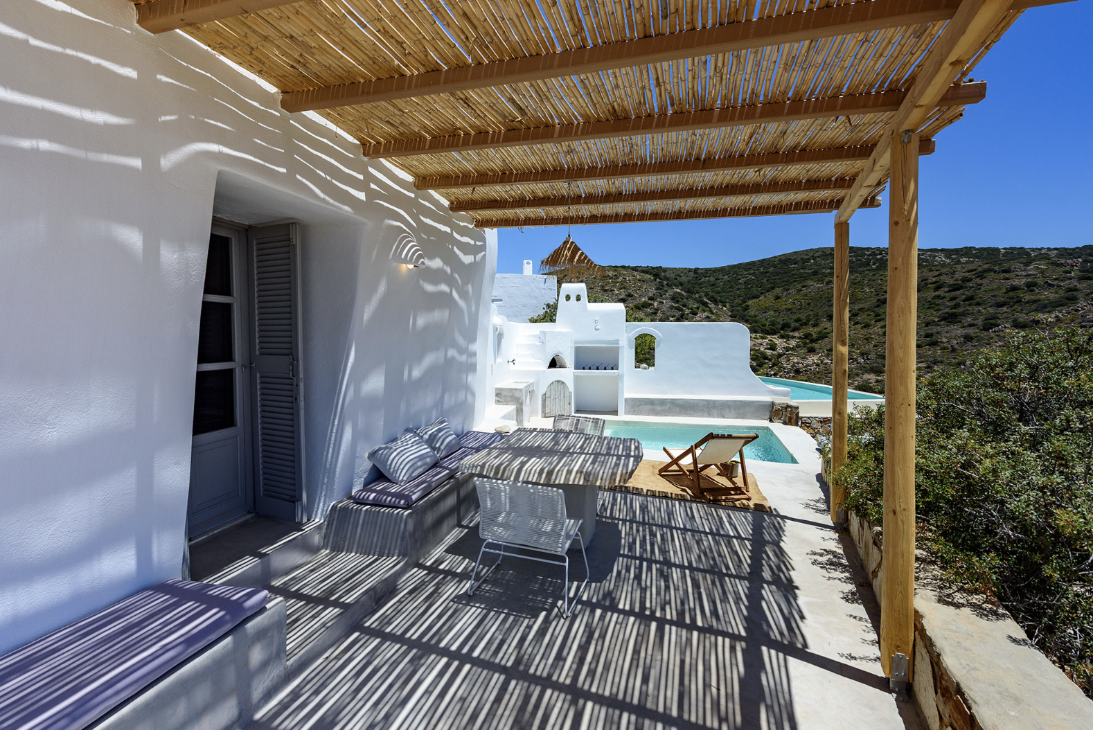 Outdoor patio with a stone table, white chair, beige bench, bean bag, and a shade structure made of bamboo and straw, overlooking a coastal landscape with hills and water under a sunny blue sky.