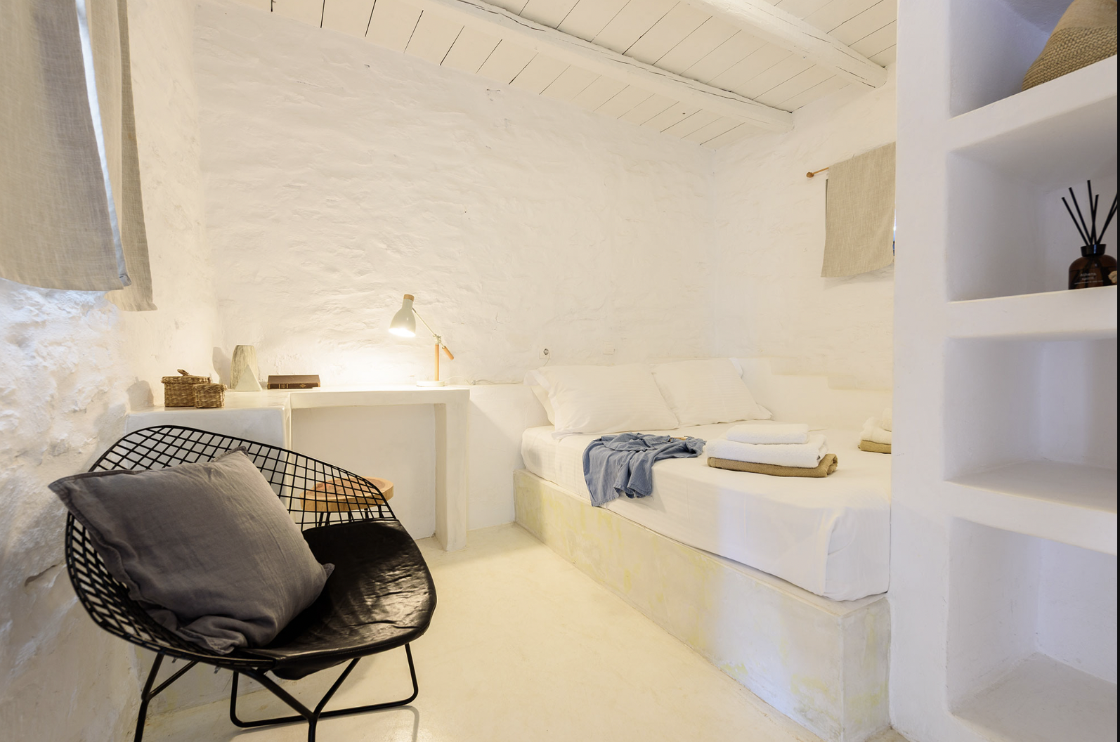A minimalist bedroom with white walls, a bed with beige pillows and a black and white blanket, and a small desk with a chair. Decor includes hanging wooden objects and woven baskets.