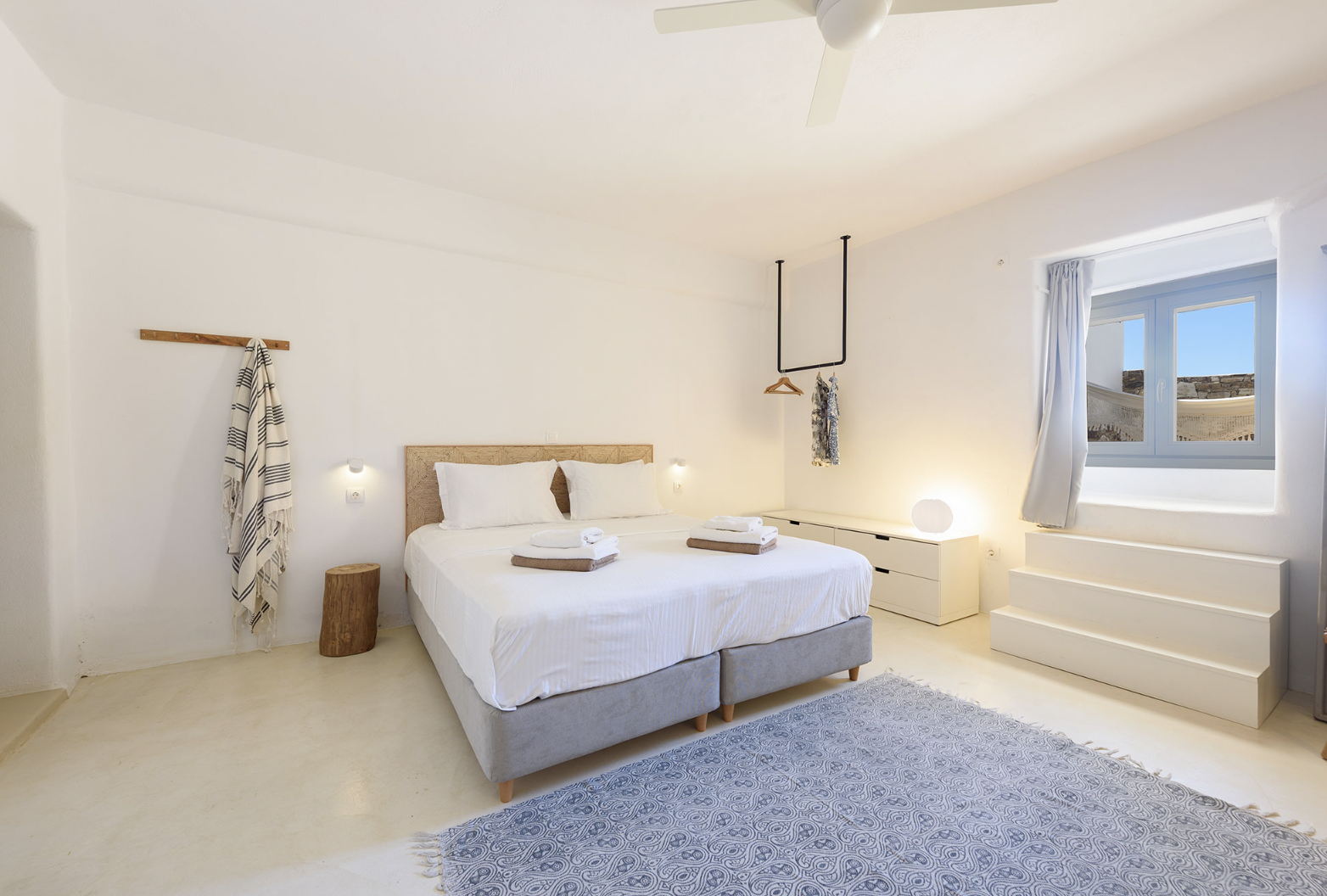 Bedroom with a bed covered in white linens, two rolled towels, a standing mirror, and large windows with gray curtains revealing a coastal view.