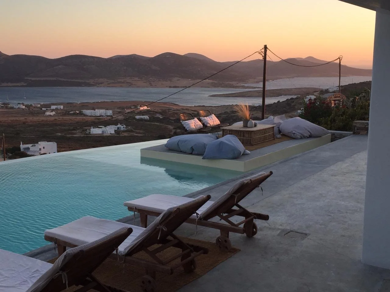 Infinity pool overlooking a coastal landscape with mountains and a body of water at sunset, with lounge chairs and cushions on a terrace.