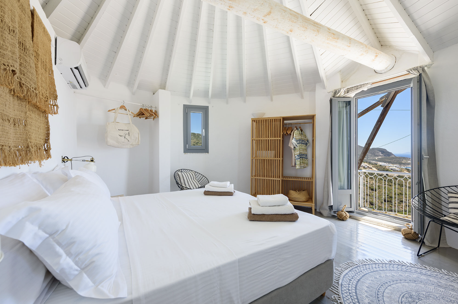 A bright, white bedroom with high, vaulted ceiling with exposed beams, a large mirror leaning against the wall, a bed with white linens and rolled towels, a small round rug, black curtains, a sliding door, and a fan in the corner.