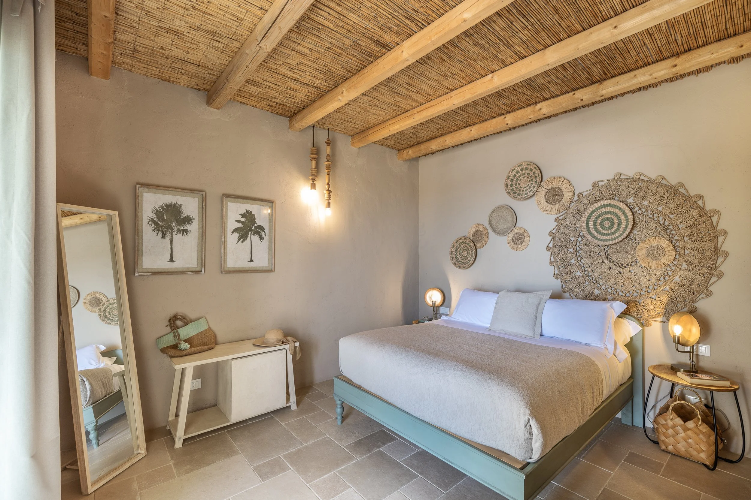 Bedroom with a bed covered in white linens, two rolled towels, a standing mirror, and large windows with gray curtains revealing a coastal view.