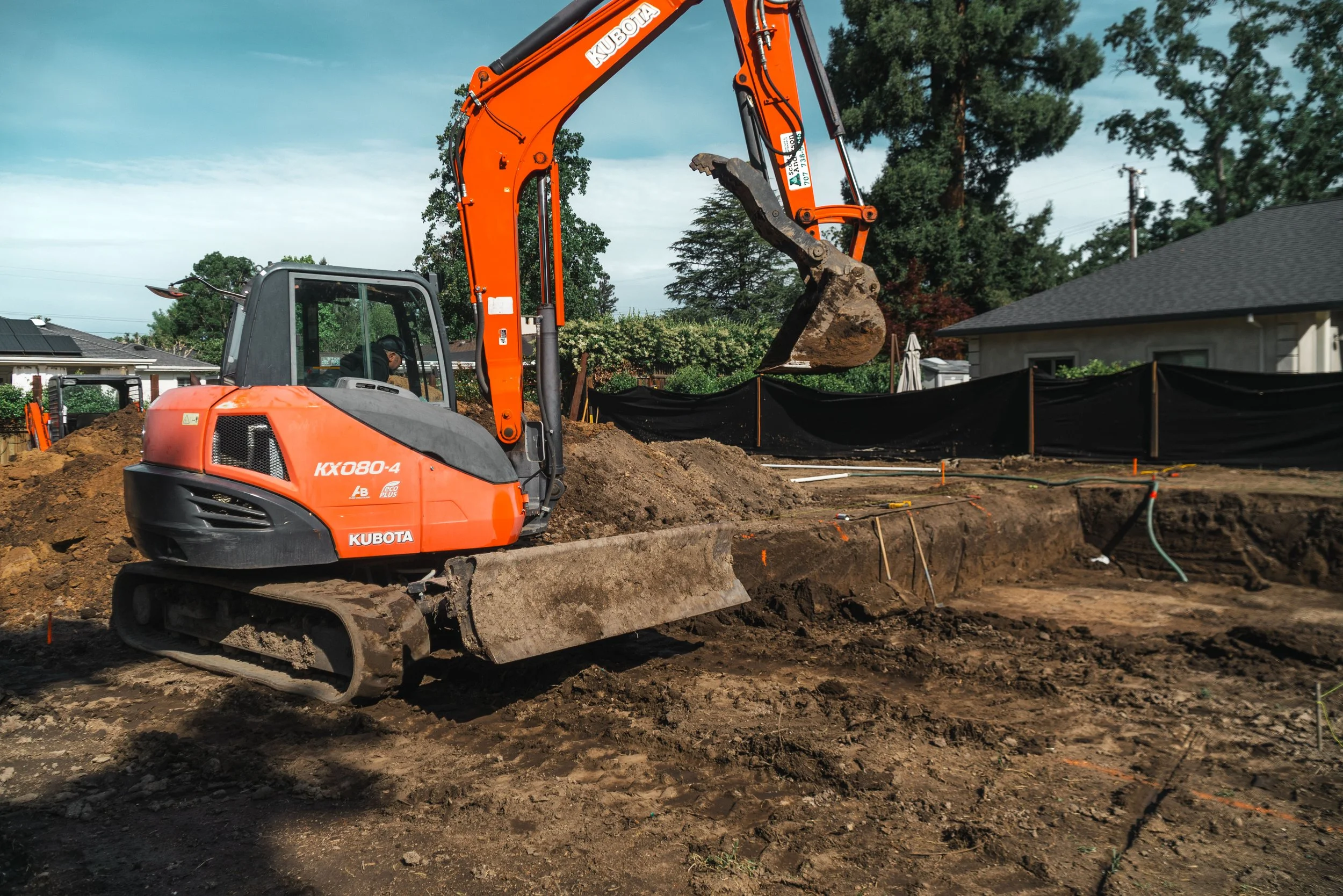 An orange Kubota mini excavator at a construction site, digging into the ground with a bucket attachment, with residential houses and trees in the background.