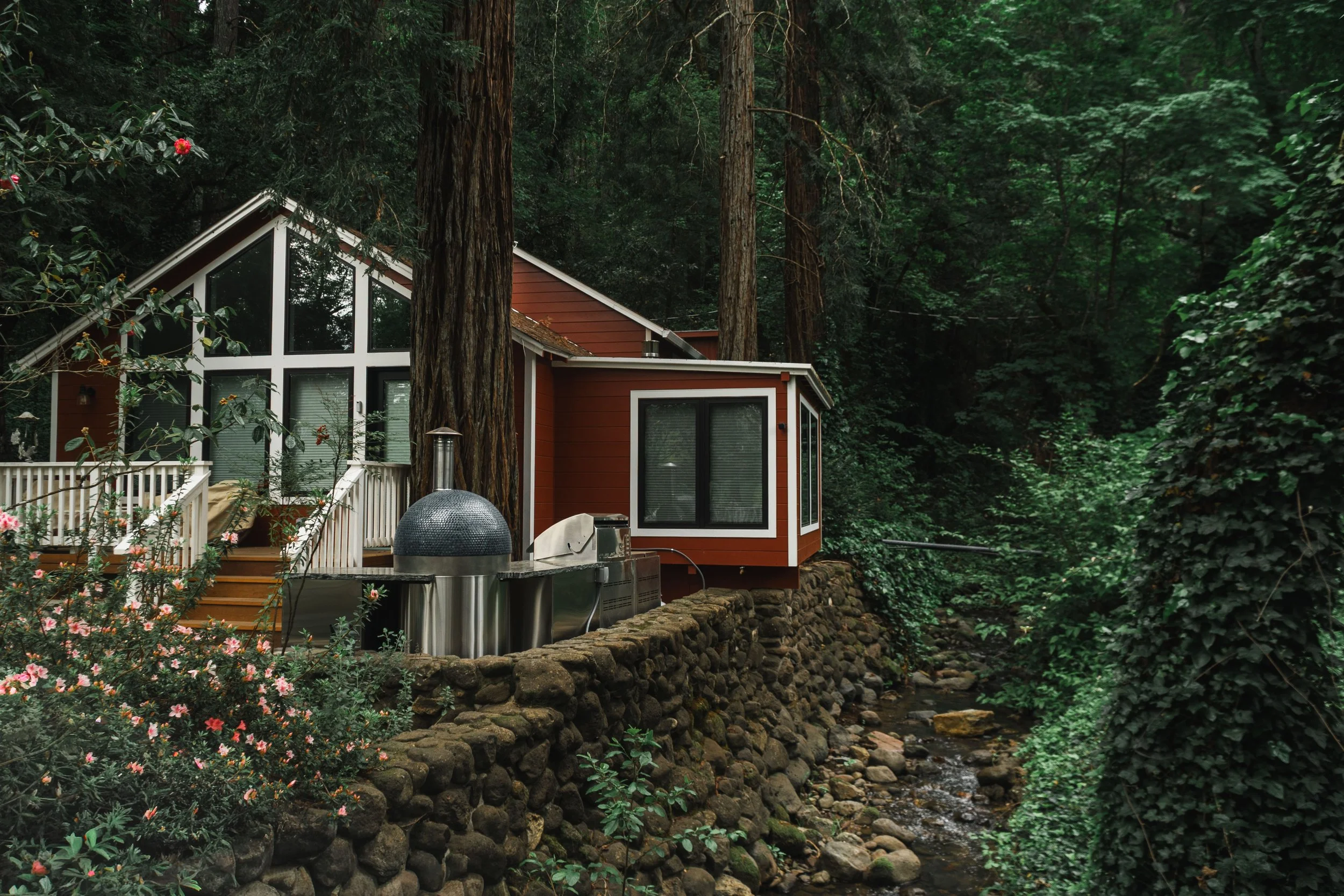 A house with large windows and a sloped roof surrounded by lush green trees and plants, built on a stone foundation over a small creek.