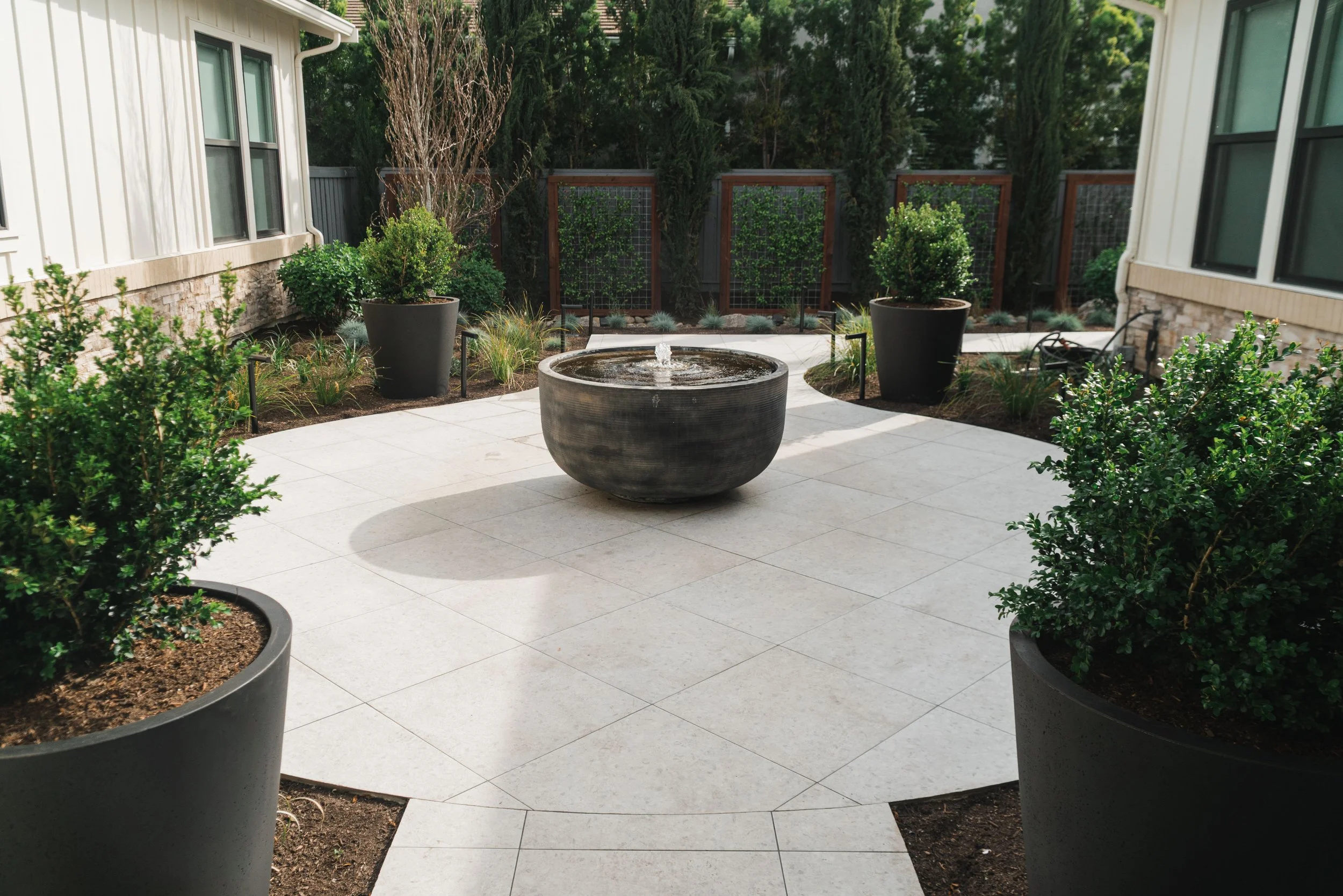 A backyard patio area with large potted bushes, a small fountain in the center, tiled flooring, and a wooden fence with greenery beyond.