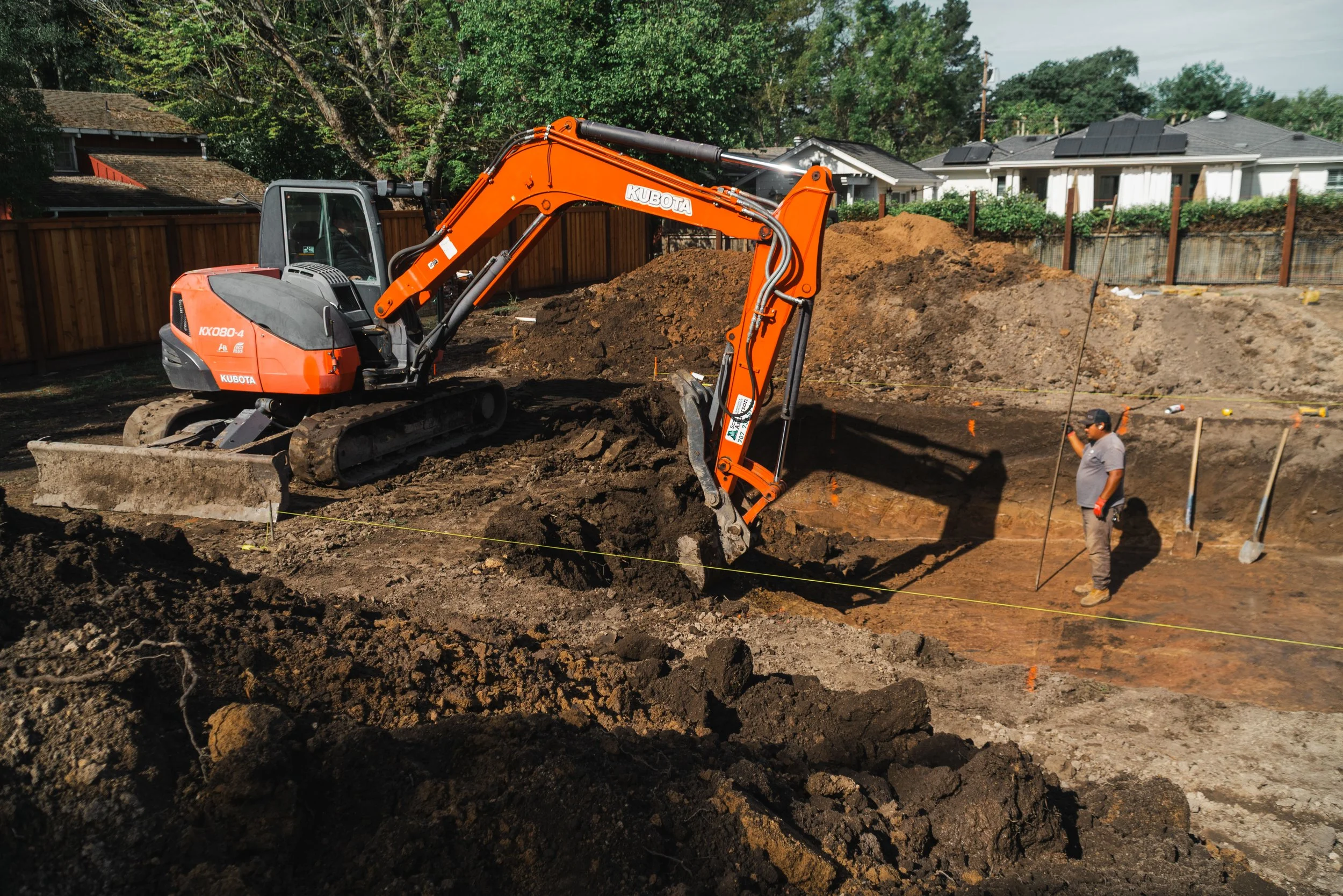 A construction site with an orange Kubota excavator digging into the earth, and two workers standing nearby with one wearing a gray shirt and black cap, surrounded by residential houses and trees.