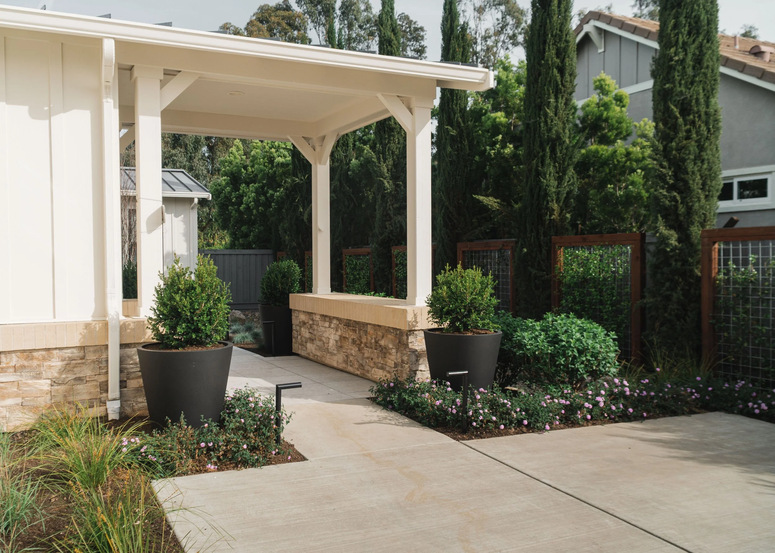 A backyard patio area with a white covered pergola, large potted plants, a stone retaining wall, and flowering bushes, surrounded by tall trees and neighboring houses.