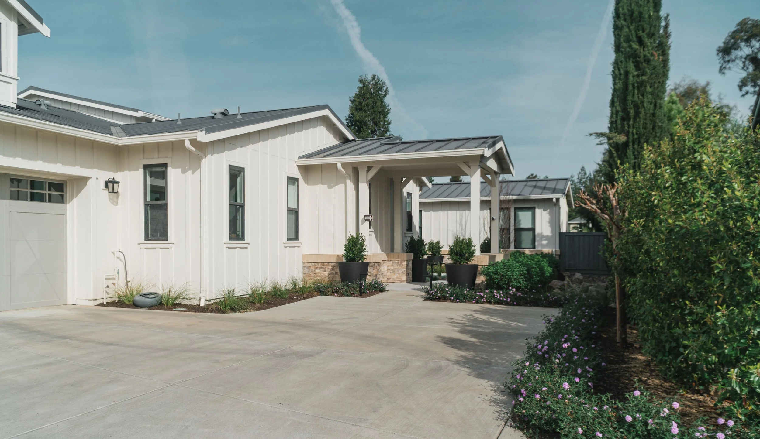 Front view of a modern white house with a covered porch, black planters with green plants, landscaped garden with flowers, and a concrete driveway under a blue sky with some clouds.