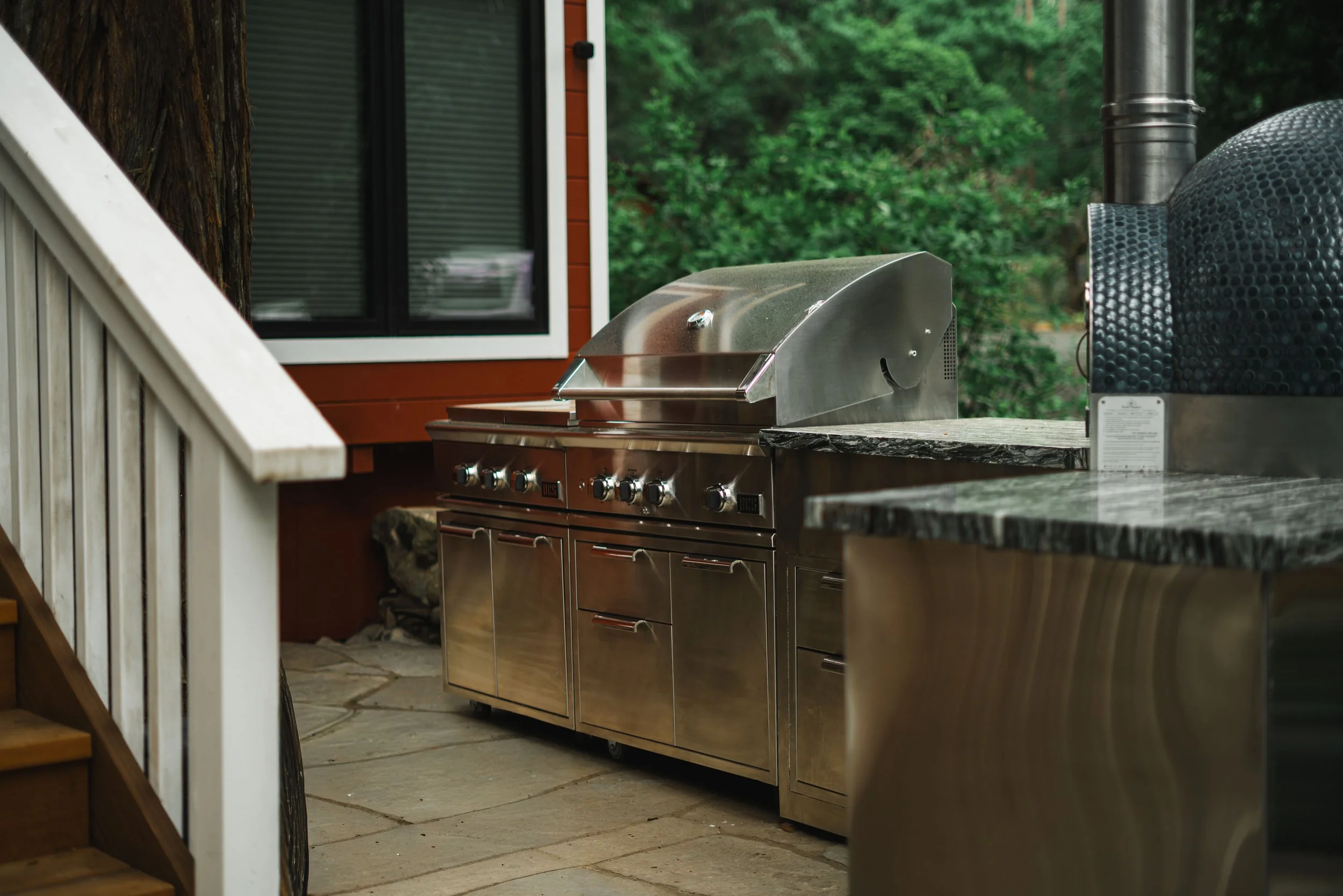 An outdoor kitchen area with a stainless steel gas grill on a stone countertop, surrounded by green trees and a wooden house wall with a window.