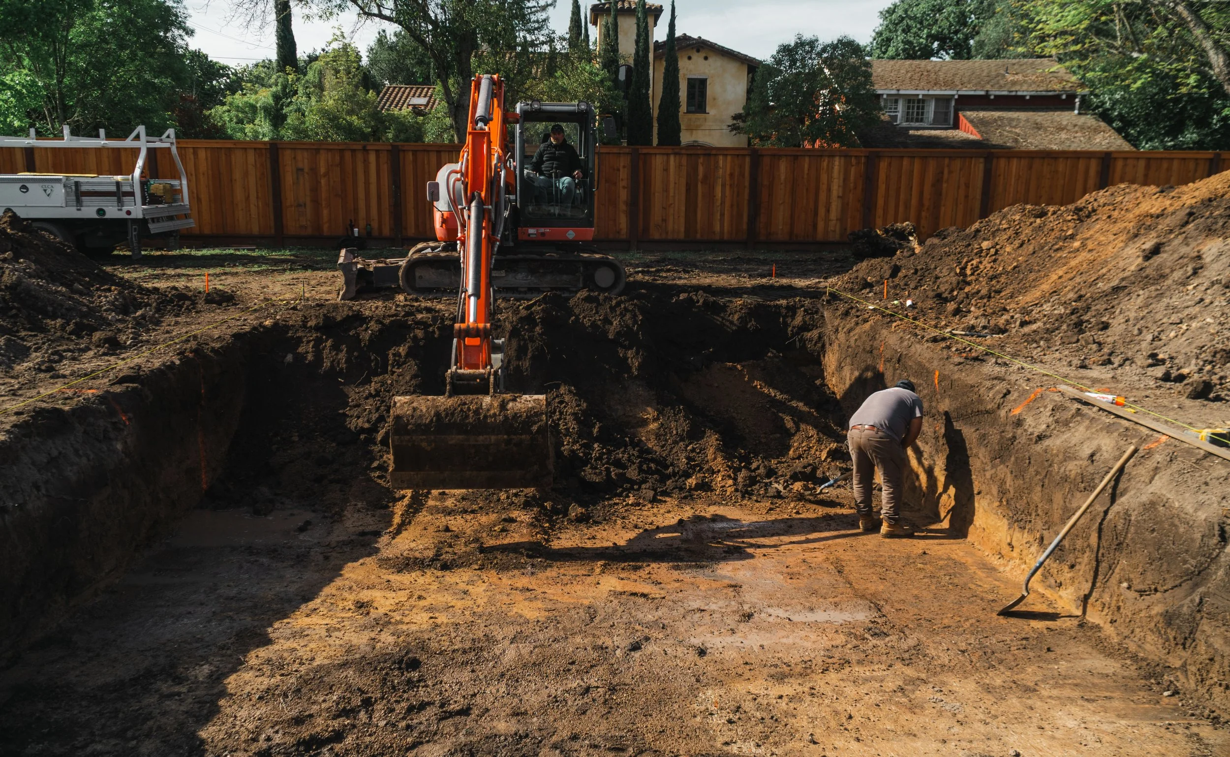 Construction site with an excavator digging a trench in a residential backyard, workers inside the trench, wooden fence, trees, and houses in the background.
