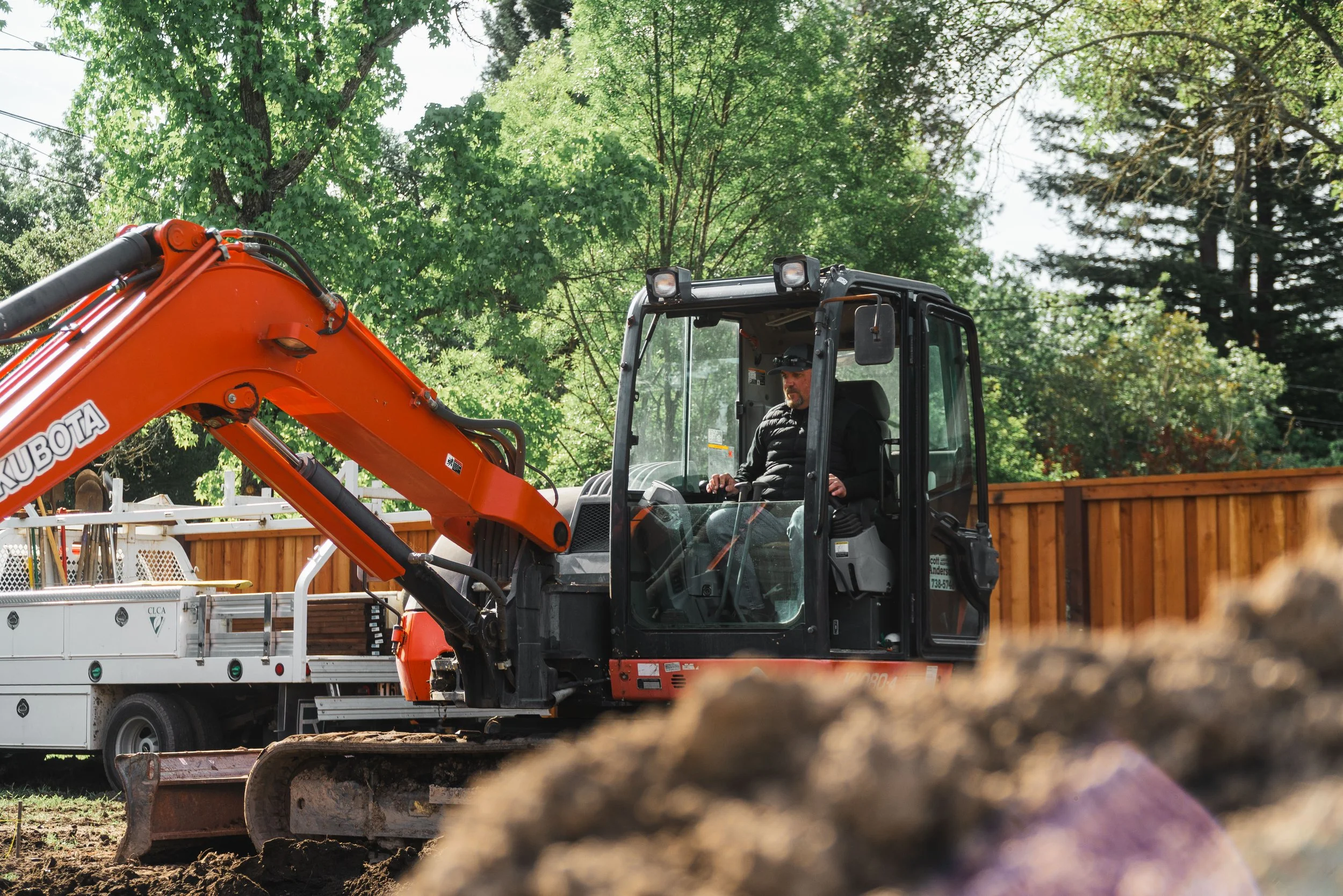 A man operating an orange Kubota excavator in a backyard, surrounded by green trees and a wooden fence, with construction equipment and dirt in the foreground.