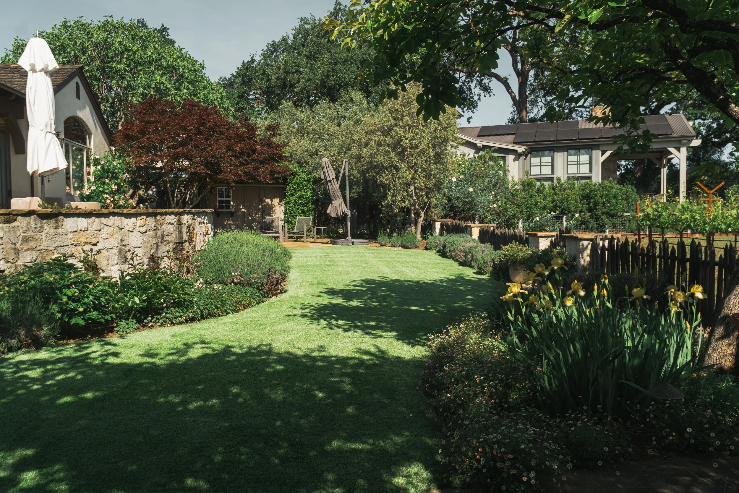 A lush backyard garden with a green lawn, various trees, and flowering plants. There is a stone wall with umbrellas and porch chairs near a house. Solar panels are visible on the roof of a neighboring building, and a fence encloses part of the garden.