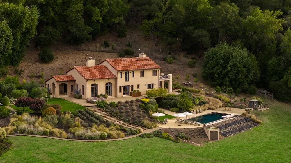 A large house with a red tile roof surrounded by a landscaped garden and a swimming pool, set in a hilly, wooded area.