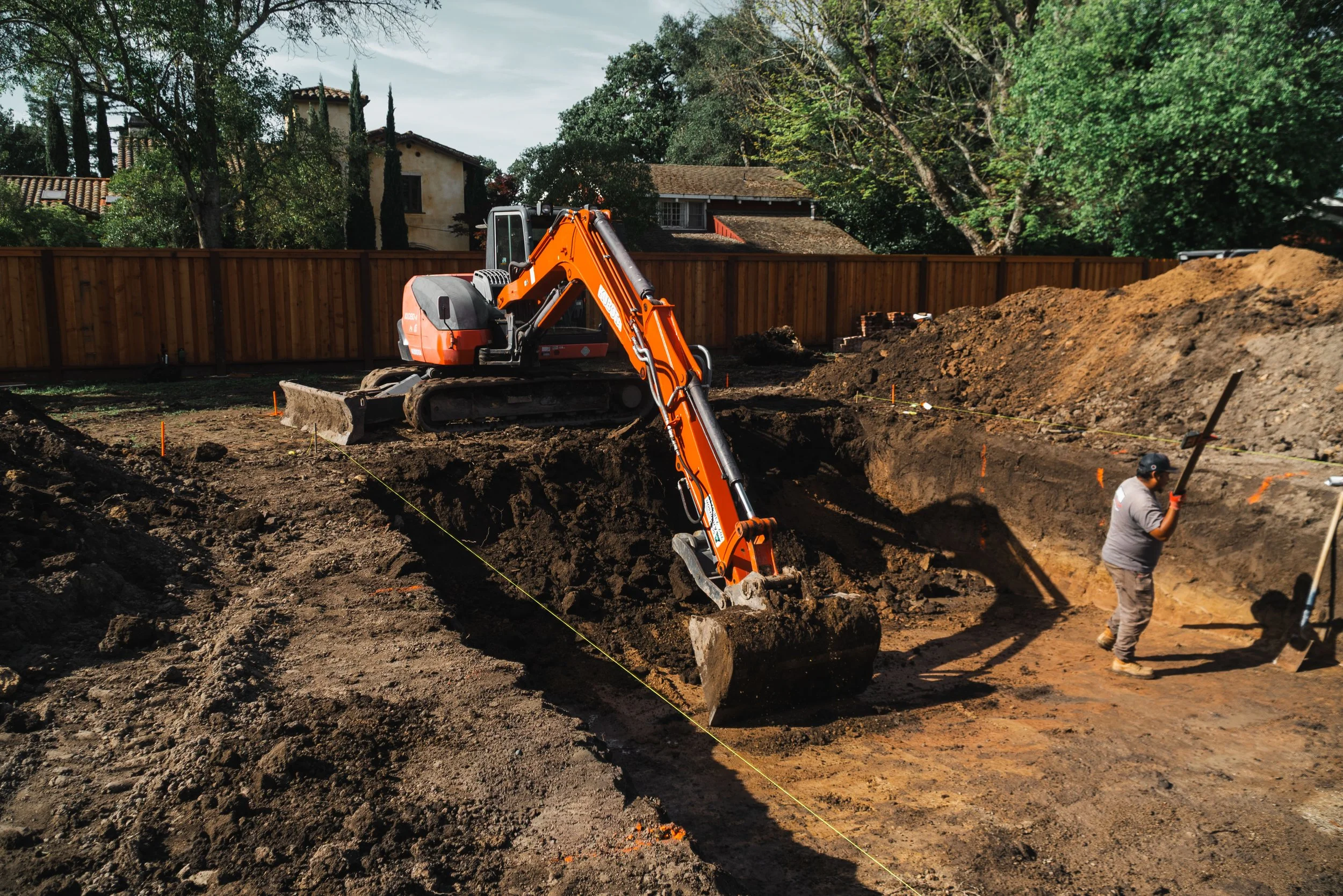 Construction site with a small orange excavator digging in the ground and workers nearby, behind a wooden fence with trees and residential houses in the background.