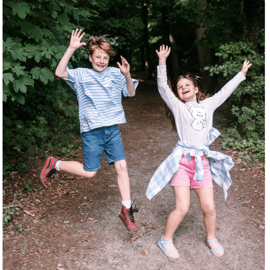 Zwei Kinder springen glücklich im Wald und entdecken spielerisch die Natur bei einer Schnitzeljagd für Kinder