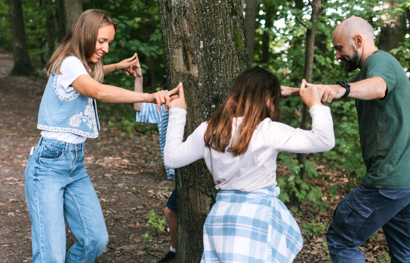 Familie spielt gemeinsam im Wald um einen Baum und erlebt eine spielerische Schnitzeljagd in der Natur
