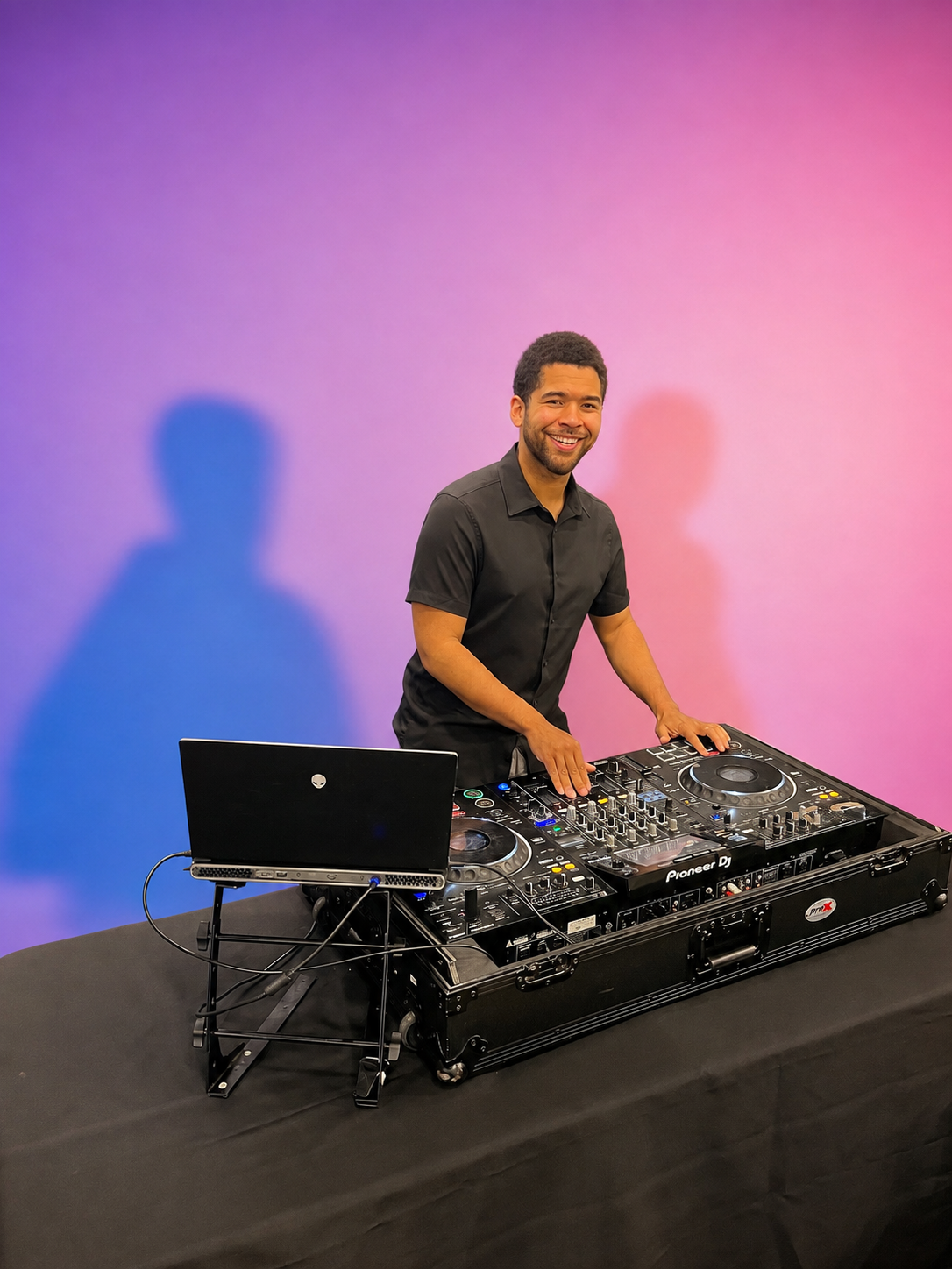 A man in a black shirt standing behind a DJ mixing console, smiling, with a laptop on a stand to his left, against a pink and purple gradient background.