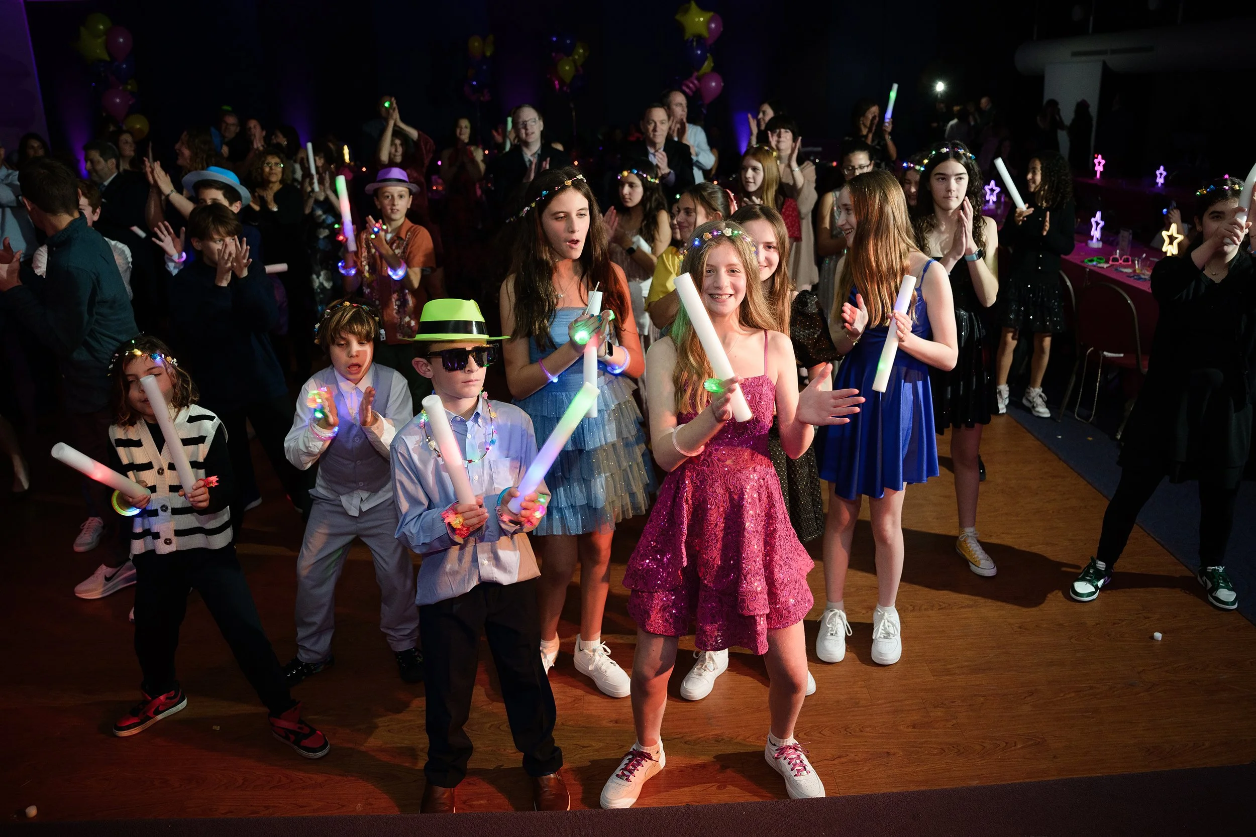 Group of children and adults dancing and celebrating at a party with glow sticks, colorful clothing, and accessories in a decorated indoor setting with balloons and party decorations.