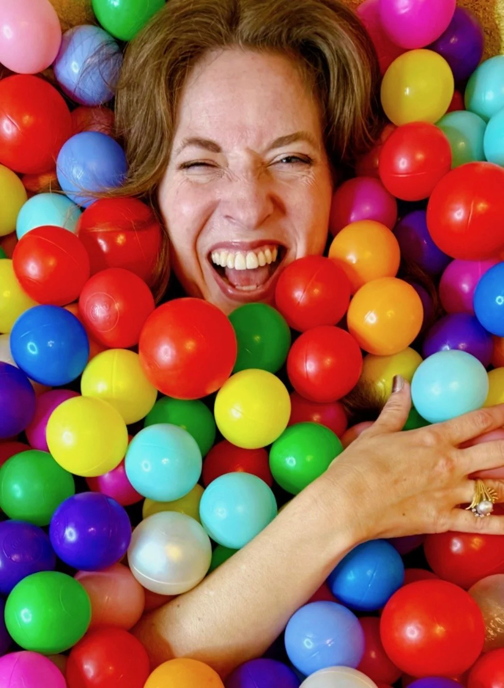 A woman with light brown hair is smiling and making a playful face while surrounded by colorful plastic balls in a ball pit.