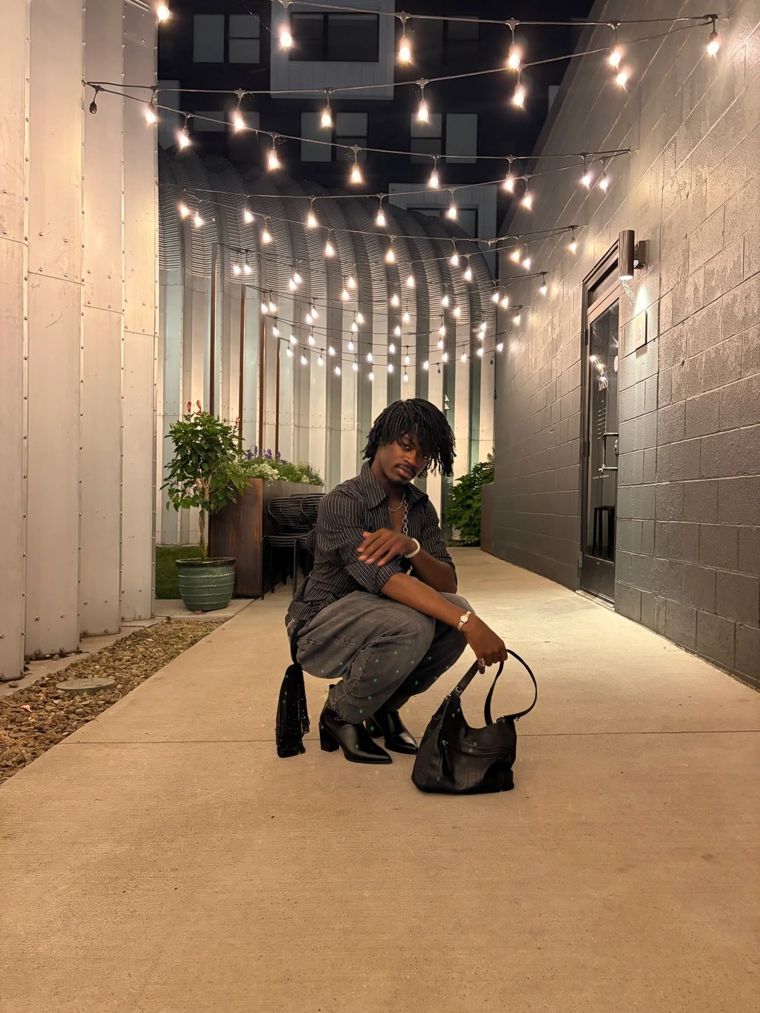 A young man with dreadlocks squatting on a sidewalk at night, holding a black bag and a pair of sunglasses. He is dressed in a striped shirt, gray pants with a pattern, and black boots. There are string lights above him, potted plants behind him, and a metal door on the right side of the image.