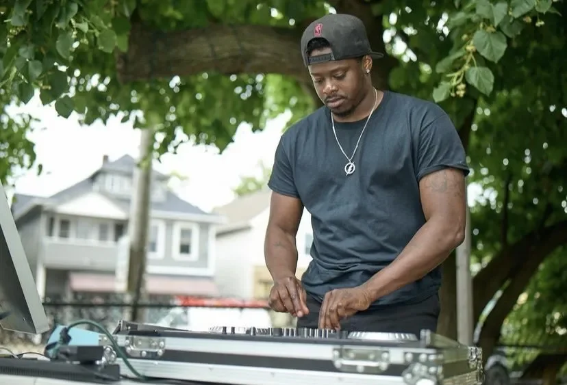 A young man wearing a black cap backwards, black t-shirt, and a peace necklace, DJing outdoors under a tree.