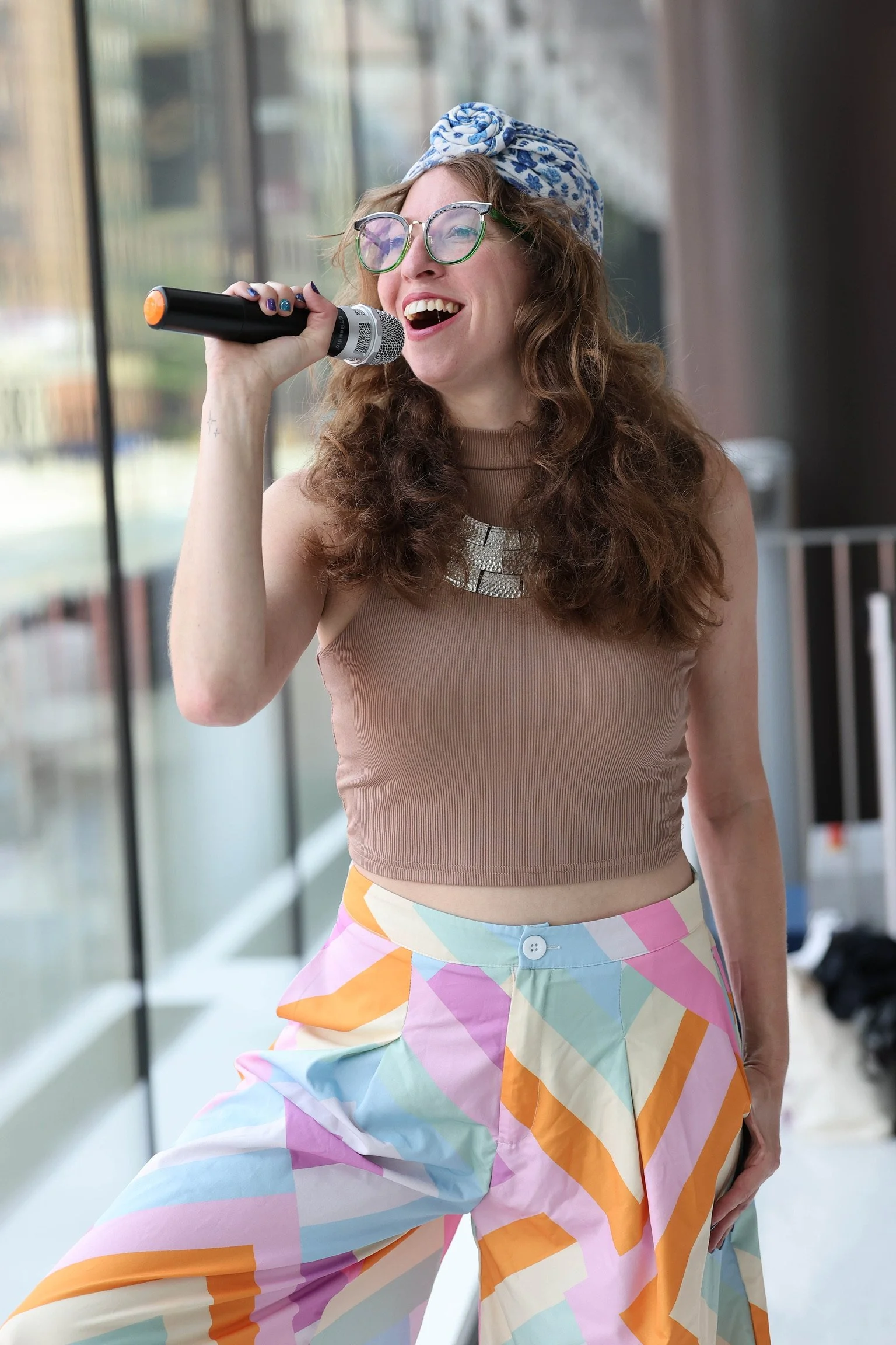A woman with curly brown hair wearing glasses, a floral headscarf, a beige sleeveless top, and colorful pants singing into a microphone indoors near a large window.
