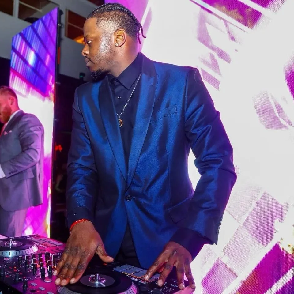 Man in a shiny blue suit DJing at an event, with large colorful screens in the background.