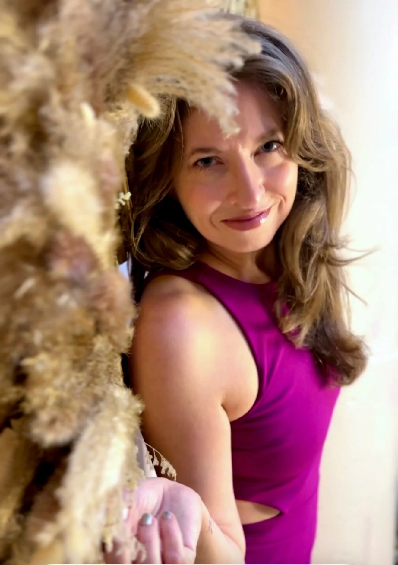 A woman with wavy brown hair and a pink sleeveless top smiling at the camera, with a furry animal partially in the foreground.