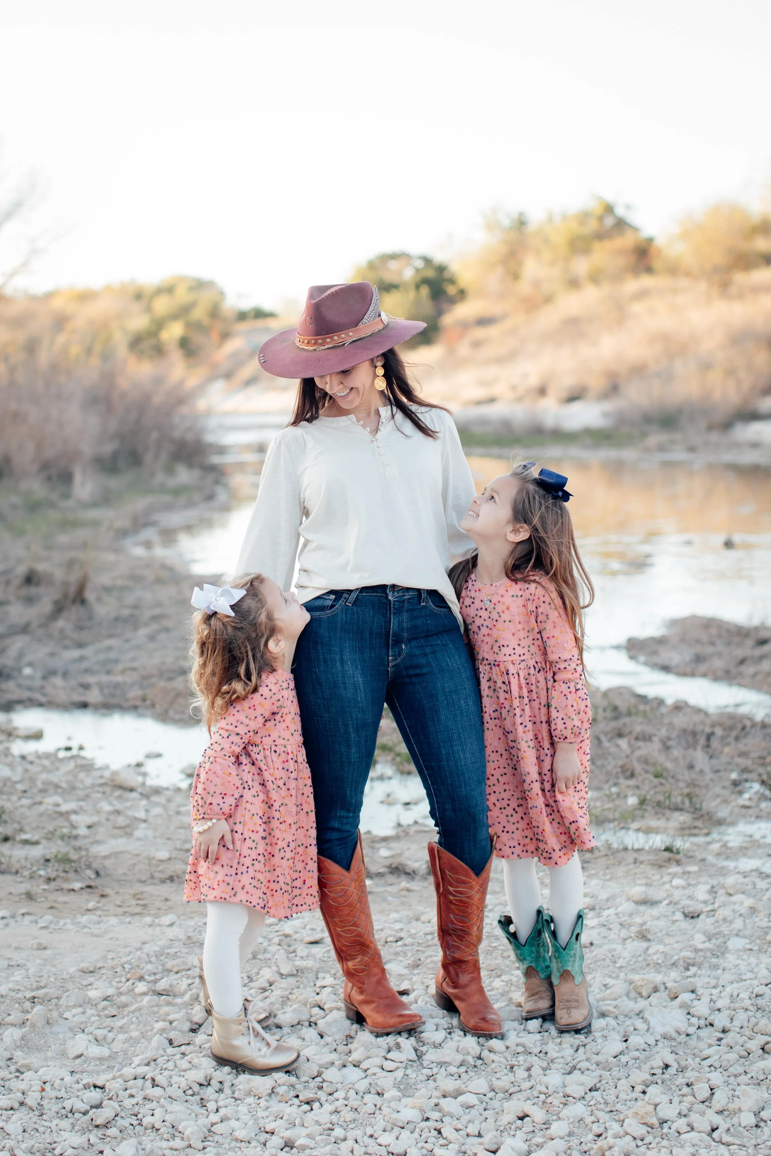 Katie Weiss, with her two daughters, standing outdoors near a river, all wearing western-style boots and dresses, smiling and looking at each other.