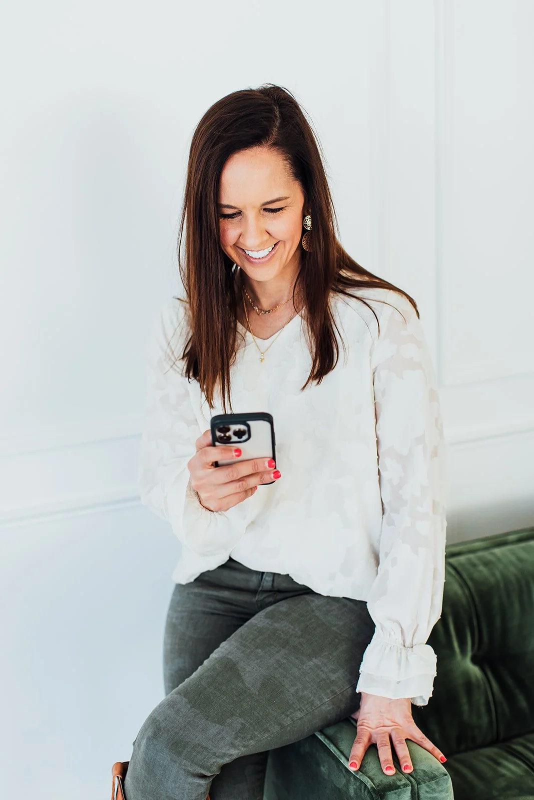 Katie Weiss, with long brown hair, wearing earrings, a white blouse, and gray pants, is sitting on a green couch, smiling as she looks at her smartphone, drafting a social media post for a client.