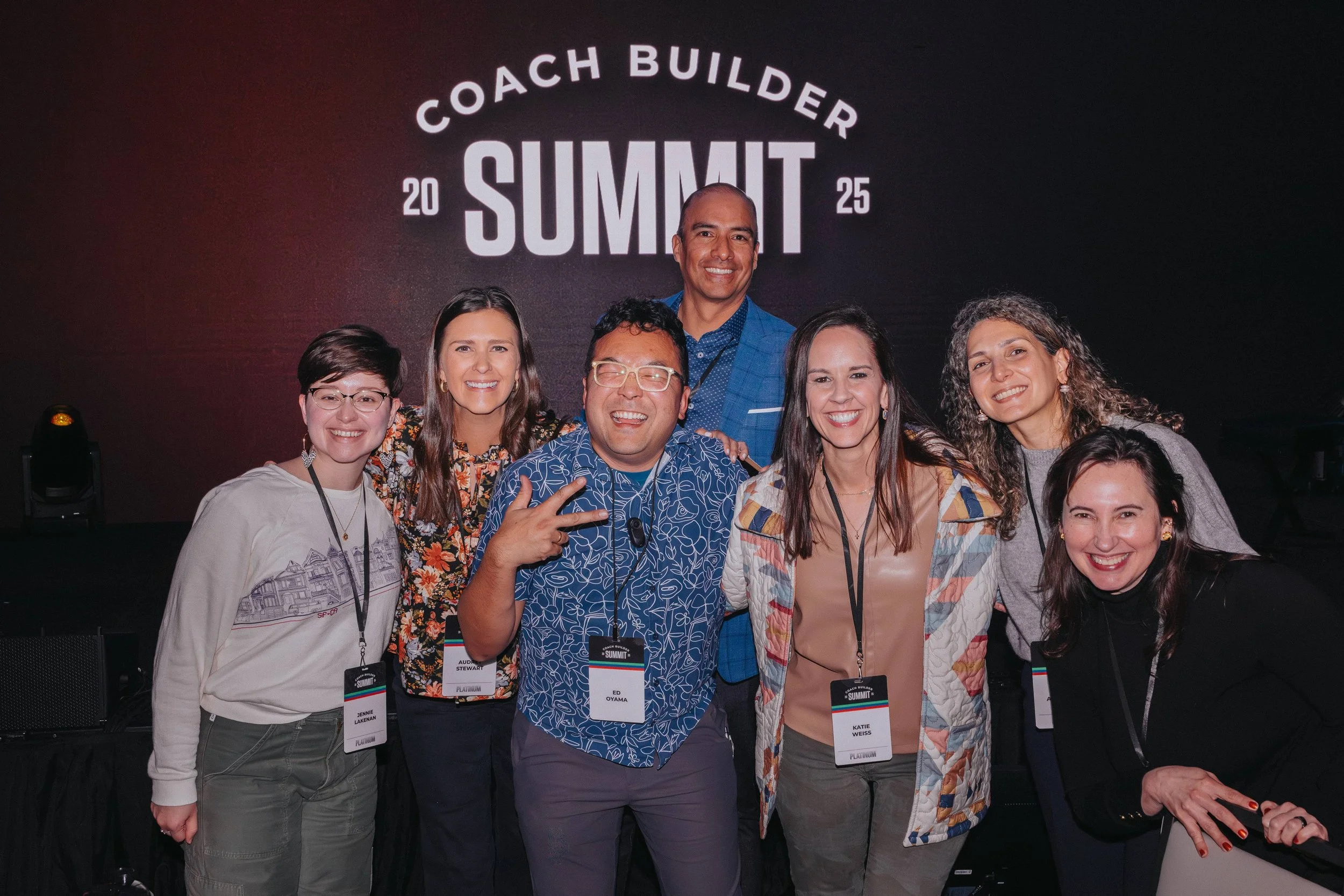 Group of seven diverse StoryBrand Guides and Business Coaches smiling and posing together at the Coach Builder Summit 2025 event, standing in front of a black backdrop with the event name and year.