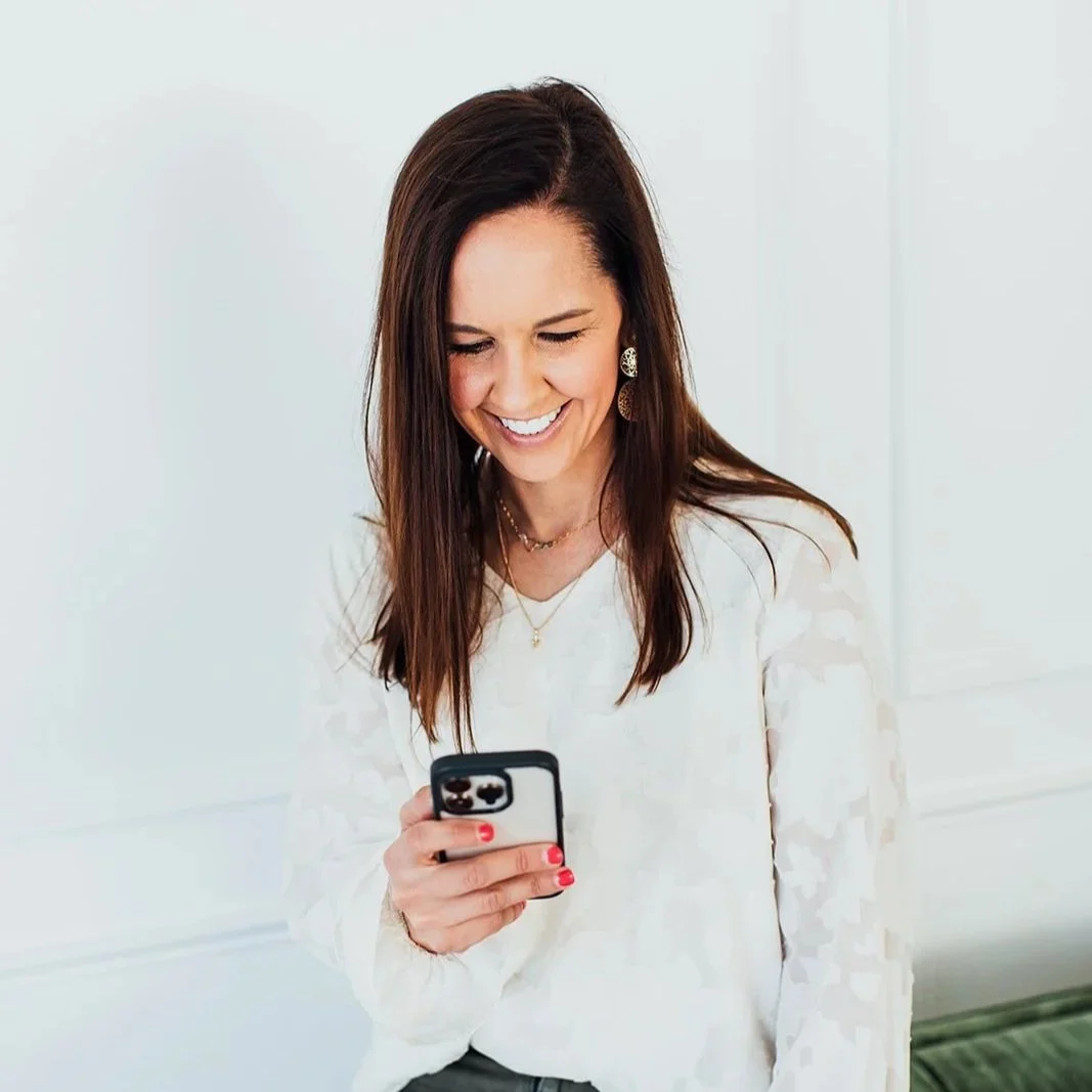 Katie Weiss, with long brown hair and jewelry, is smiling while looking at her phone, creating social content for her client.