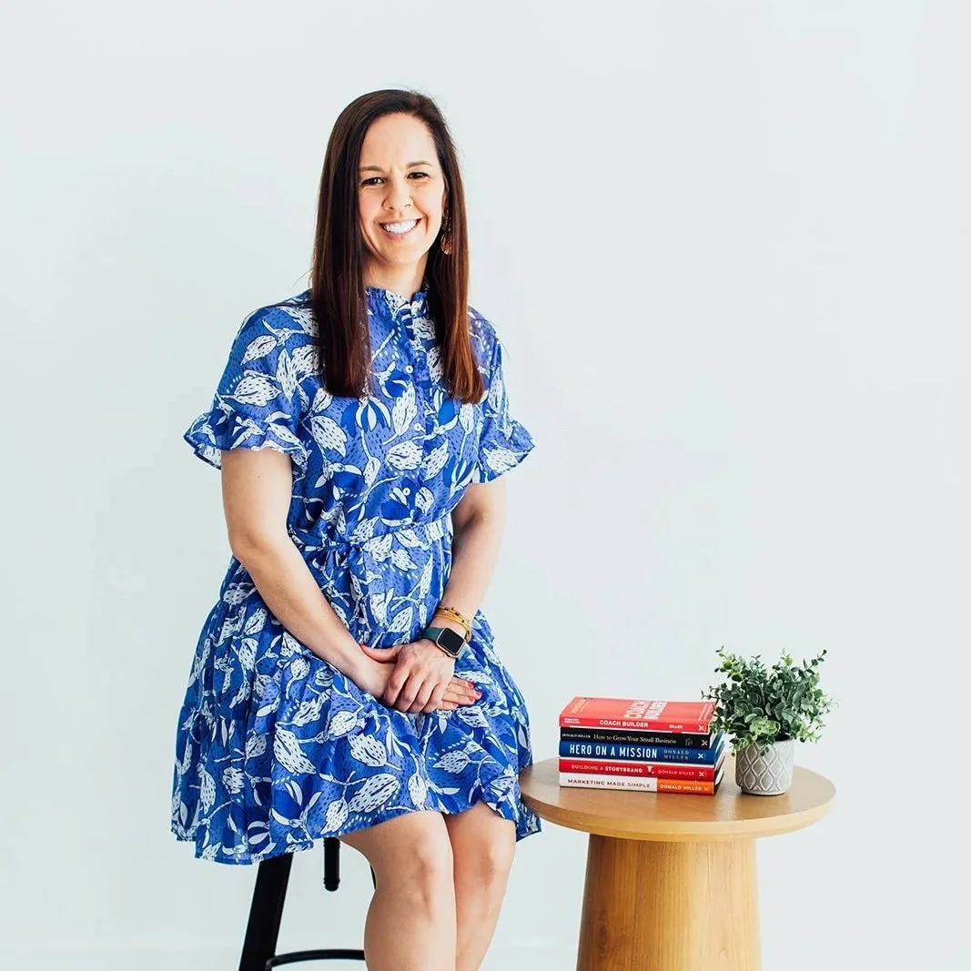 Katie Weiss is sitting on a stool, wearing a blue floral dress, smiling, with a stack of Donald Miller's business books: CoachBuilder, Business, and a small potted plant on a round wooden table next to her, against a plain light-colored background.