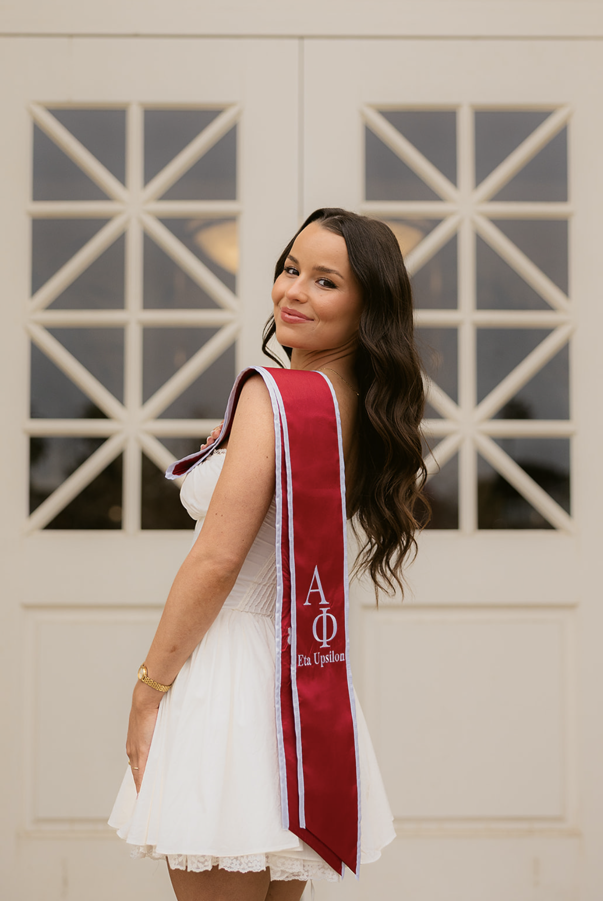 A young woman with long, wavy brown hair, wearing a sleeveless white dress with lace trim, stands in front of a white paneled door with glass windows. She has a maroon sash with Greek letters and the words 'Eta Upsilon,' smiling and looking over her