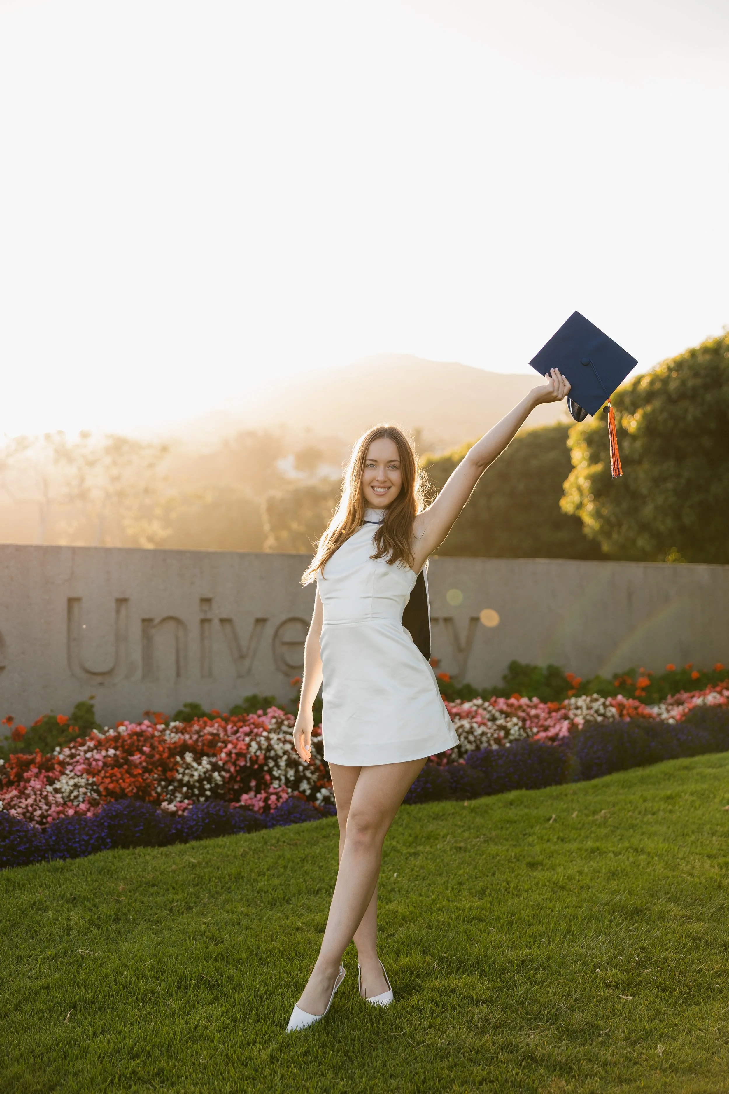 A young woman in a white dress holding a graduation cap, celebrating outdoors on a sunny day with flowers and trees in the background.